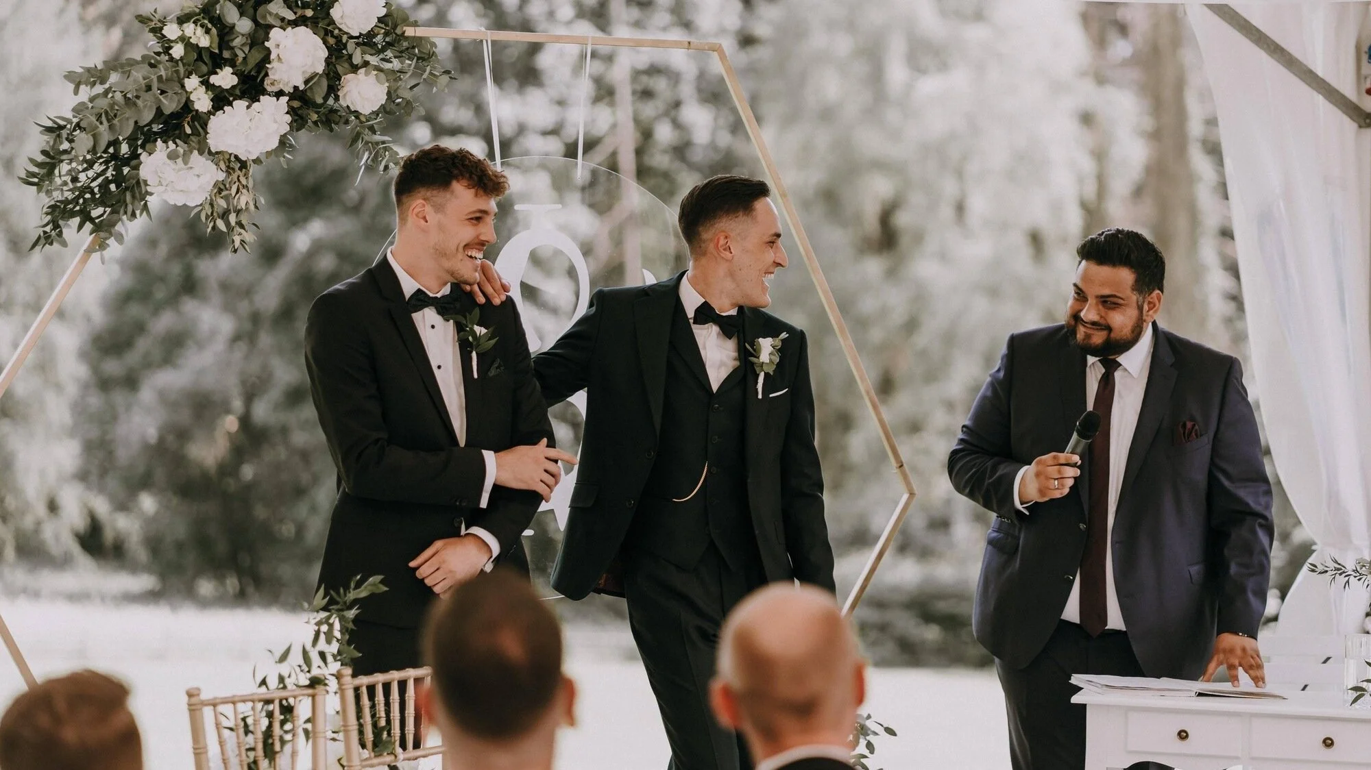 Der freie Trauredner Pedi mit einem Bräutigam und seinem Trauzeugen in dunklen Anzügen bei einer Hochzeit, der eine hält ein Mikrofon, im Hintergrund eine Blumen- und Baumdekoration im Freien.