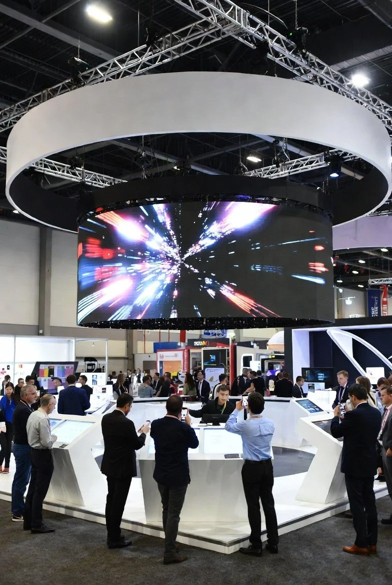 People gathered around technology displays at a trade show. There is a large, circular LED screen overhead showing a colorful, dynamic digital graphic.