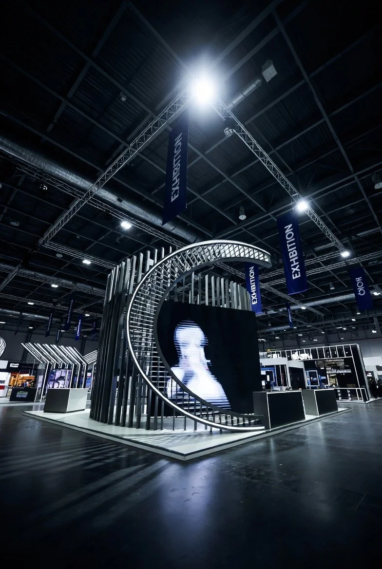 Exhibition booth with metallic curved structures and a large LED screen at an indoor event.