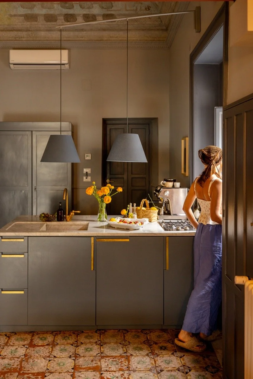 A woman standing in a kitchen near an open doorway with sunlight streaming in, with a gray kitchen counter and two gray hanging lamps above, a vase of yellow flowers, and various kitchen items.