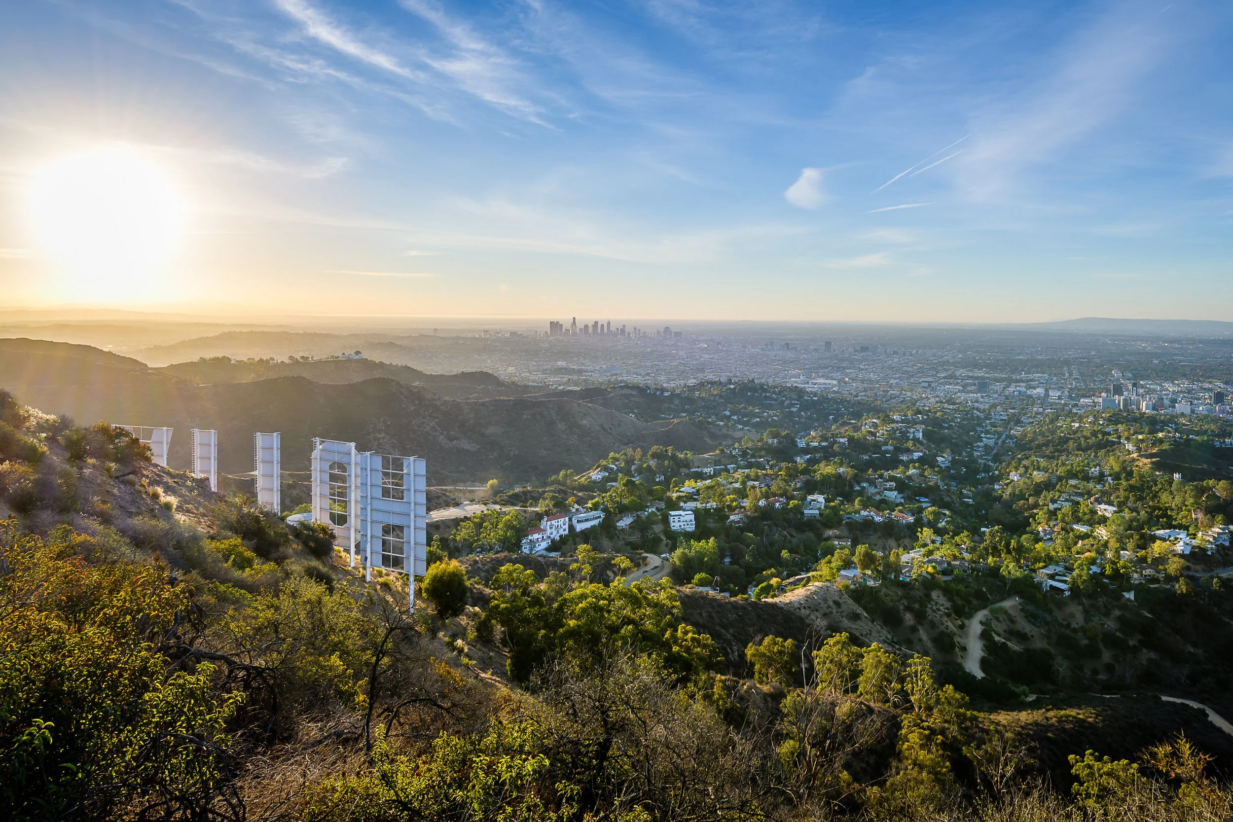 View of Los Angeles city skyline from a hillside during sunset with clear blue sky and some clouds.