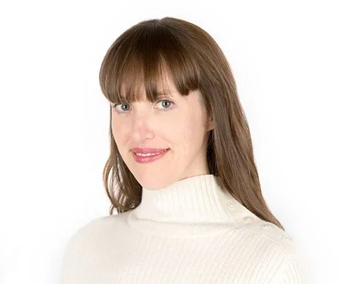 A woman with straight brown hair, and bangs, wearing a white turtleneck sweater, smiling against a plain white background.