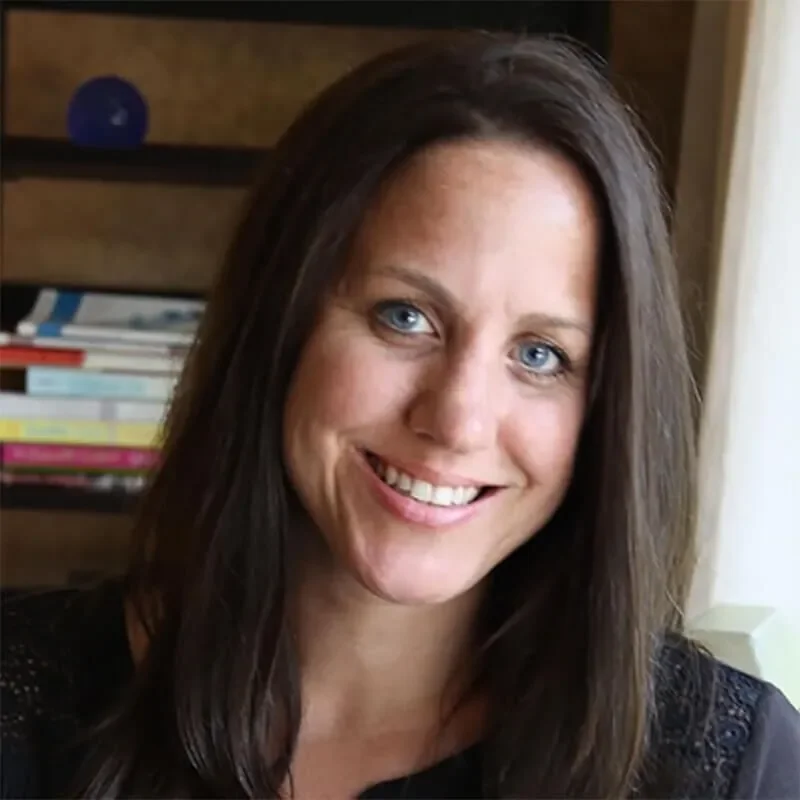 A woman with long dark hair and blue eyes smiling in front of a bookshelf.