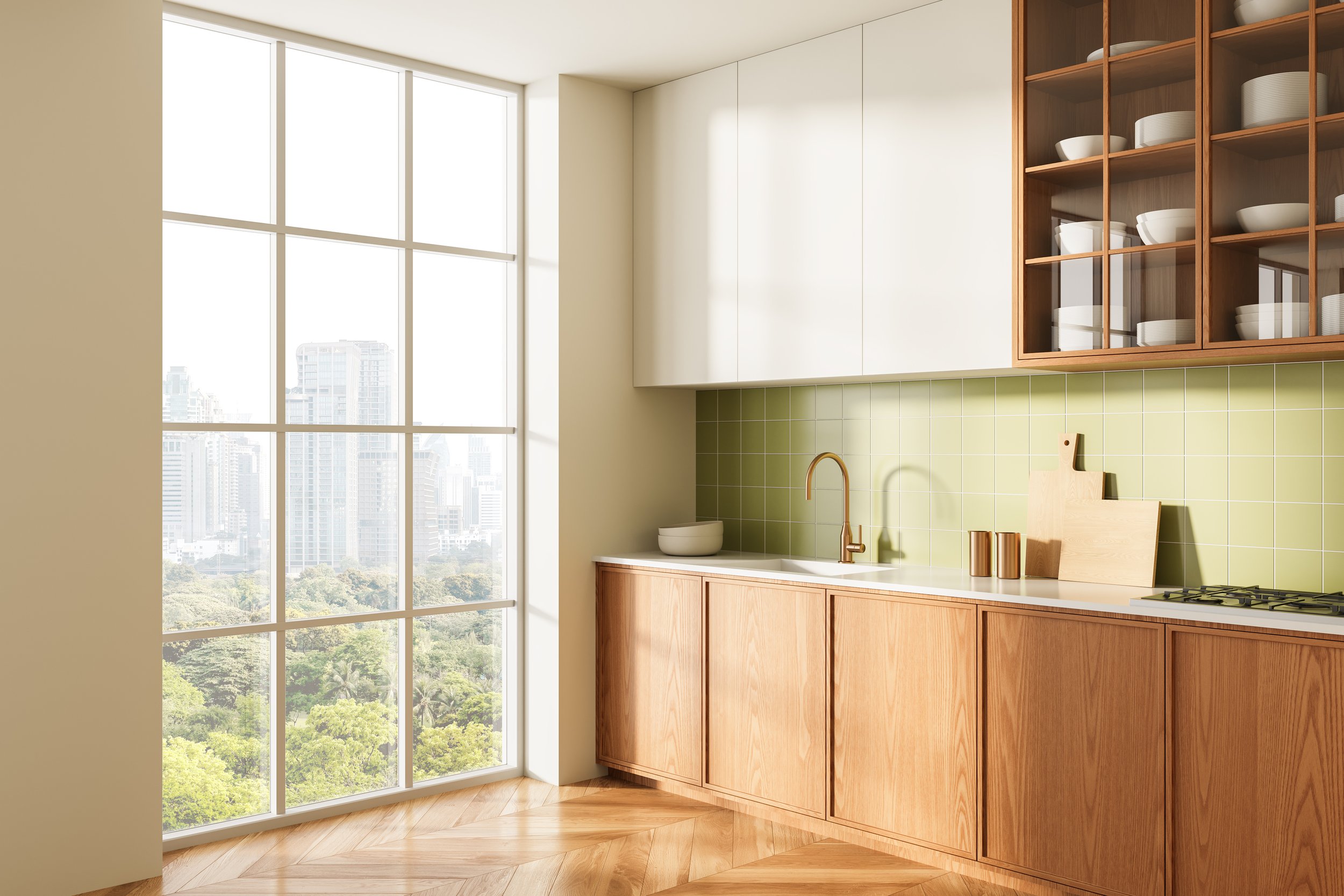 A modern kitchen with large floor-to-ceiling windows showing a cityscape and greenery outside. The kitchen features wooden cabinets, a white countertop, green tile backsplash, and open wooden shelving with white and gray dishes. A gold faucet and a small white bowl are on the countertop.