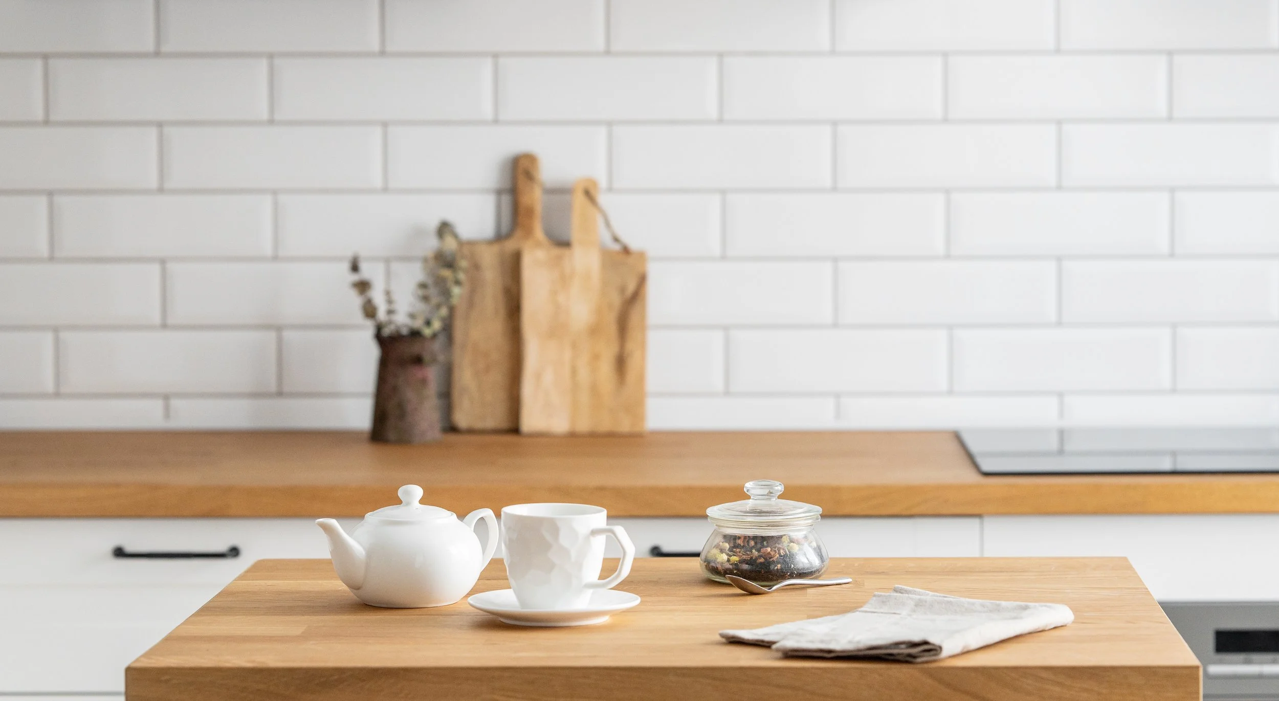 A kitchen countertop with a white teapot, a teacup, a glass jar of tea, and a spoon. In the background, there are white subway tiles on the wall and wooden cutting boards with a small plant.