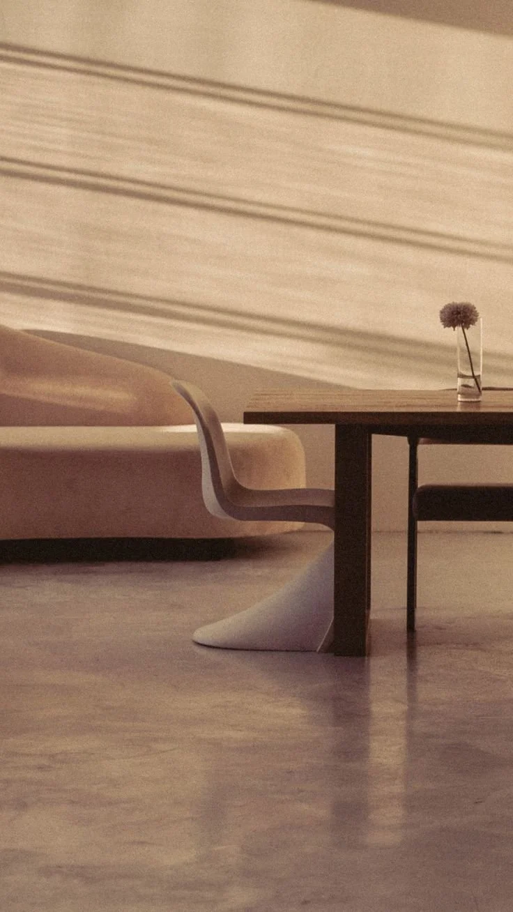 A minimalist interior scene with a wooden table, a beige modern chair, and a pinkish sofa. A small vase with a single flower sits on the table. The background features a lightly striped wall.