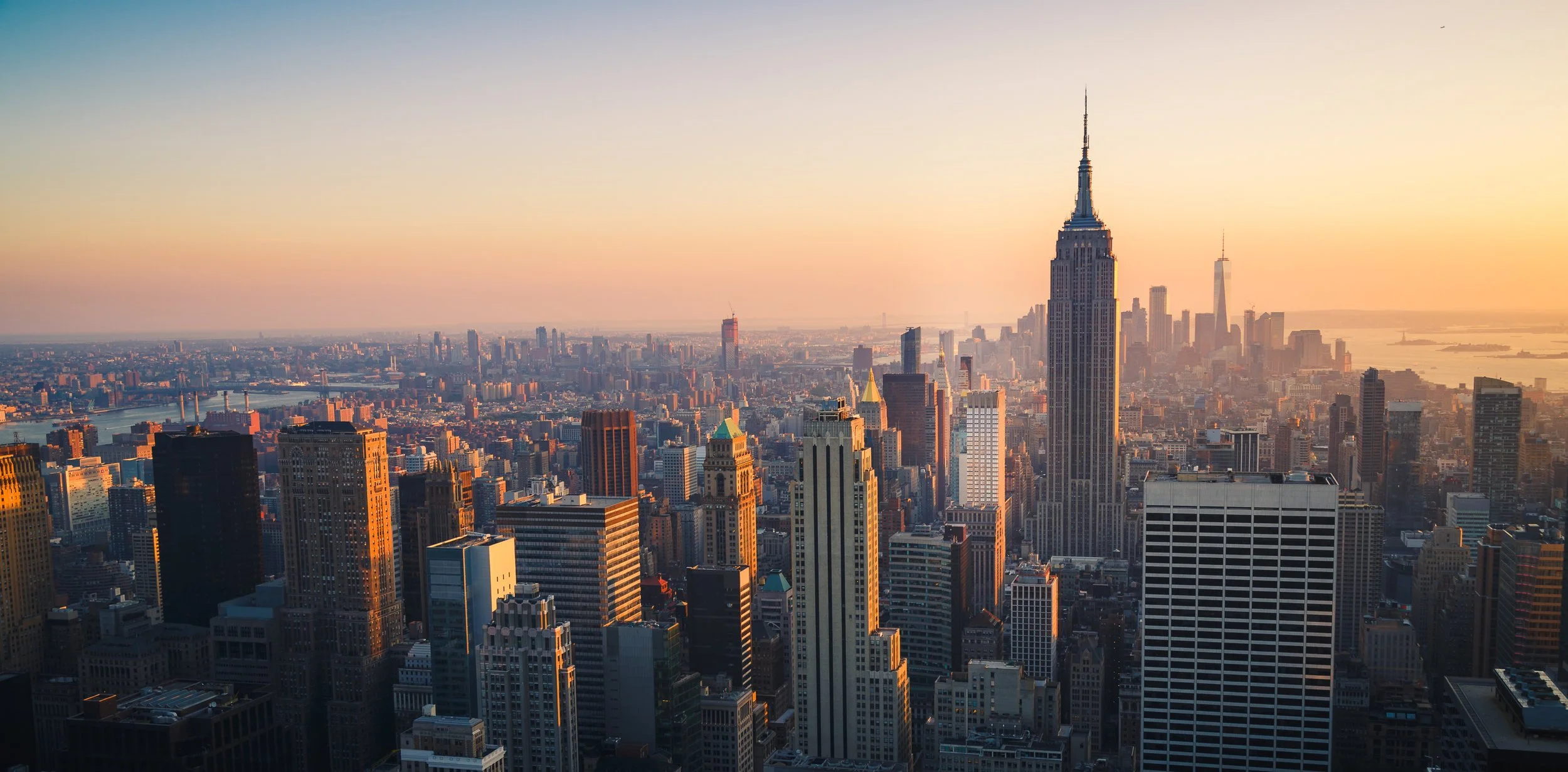 Tall skyscrapers of Manhattan skyline in New York City at sunset, with the Empire State Building prominently visible and other iconic buildings in the background.