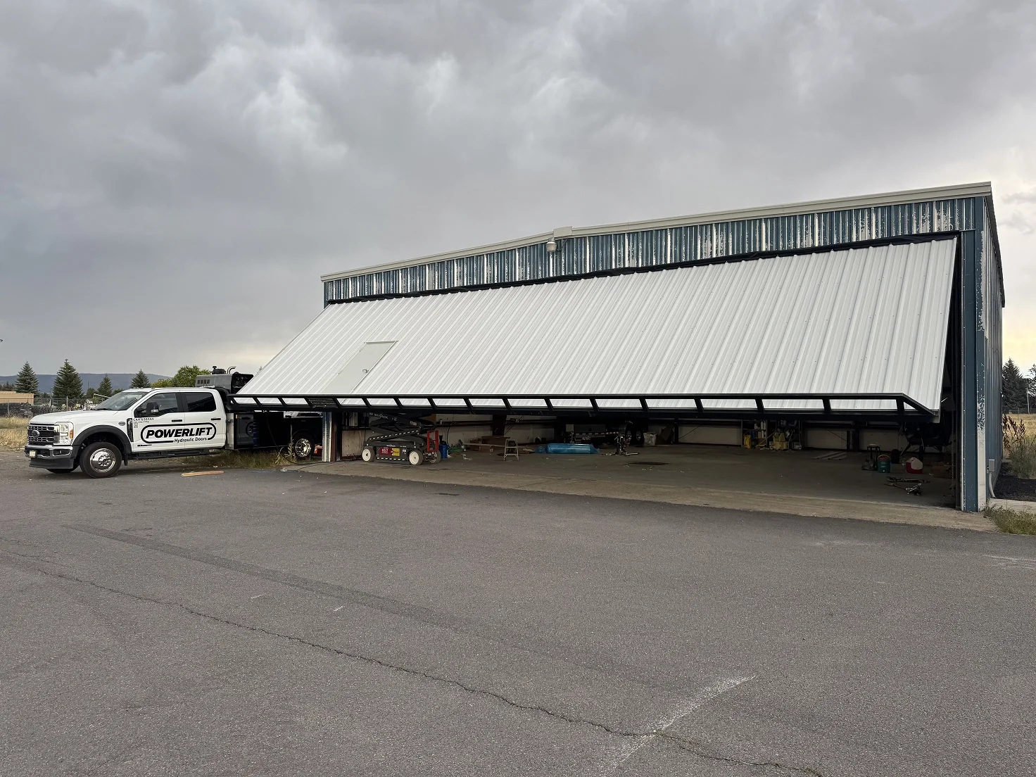A large industrial garage with a steel sliding door; part of the door is open, revealing tools and equipment inside. A white pickup truck with black lettering 'Powerlift' is parked outside.