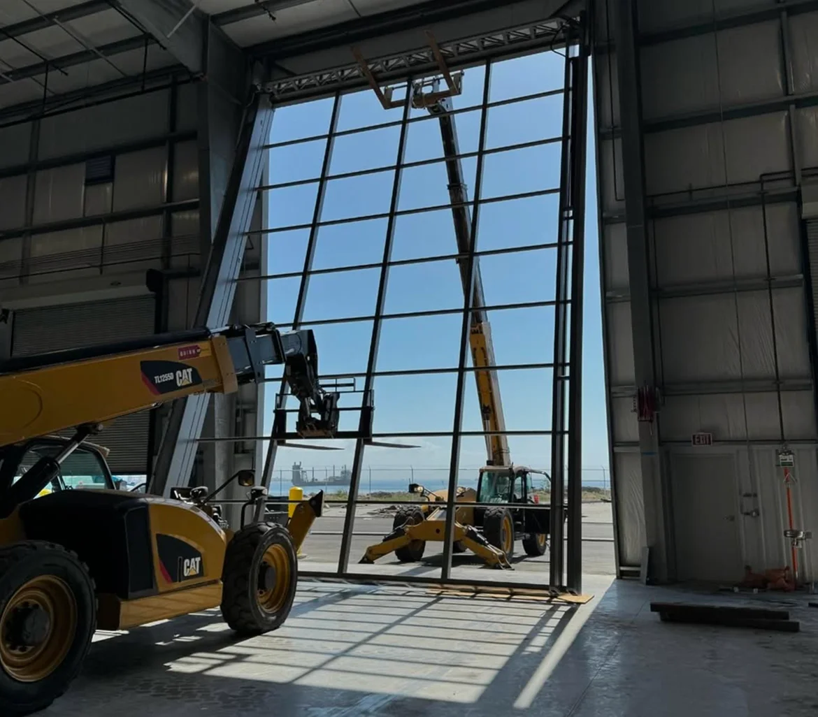 Construction workers use cranes and a forklift to install large glass panels in an airport hangar or warehouse window.