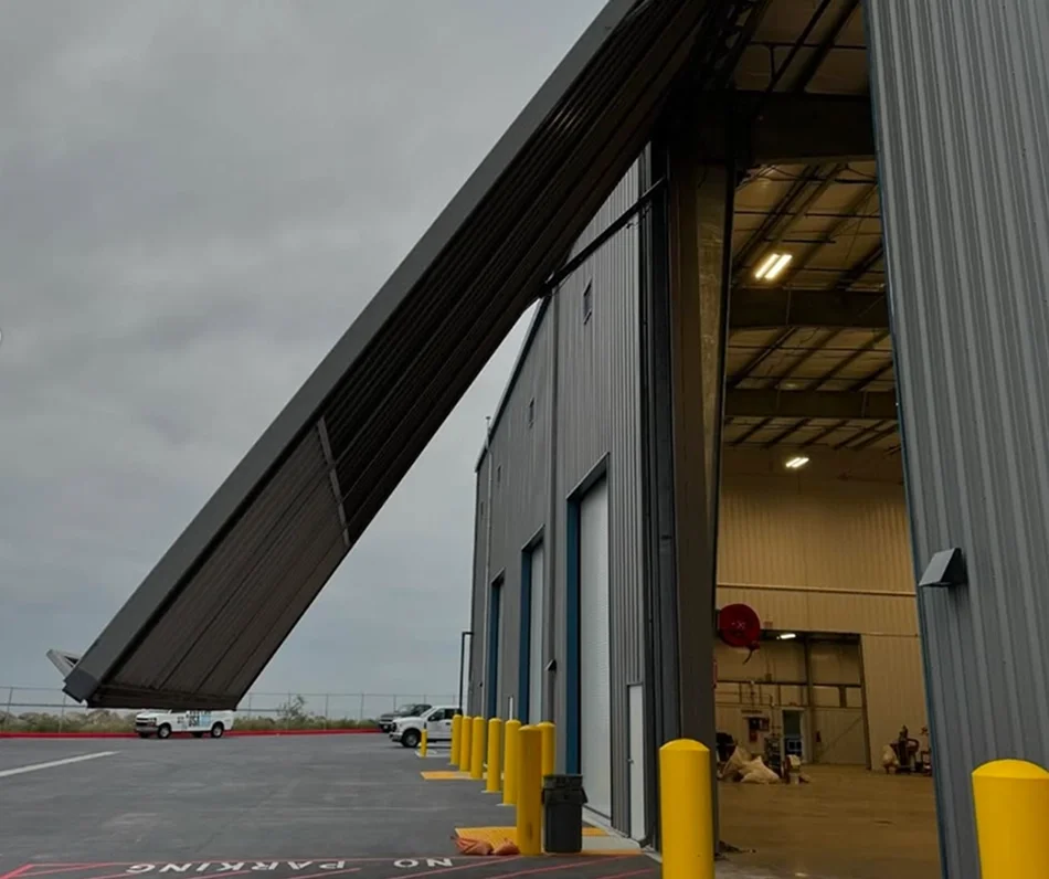 A large industrial storage building with a partially open roll-up door, showing the interior. A long metal ramp extends from the roof to the ground, possibly for loading or maintenance. Several yellow safety bollards are positioned in front of the building. The parking lot is mostly empty with a couple of trucks and vehicles.