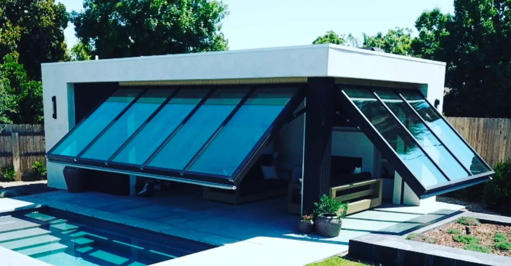 Modern white house with large glass windows and a small pool in the front yard, surrounded by a wooden fence and trees.