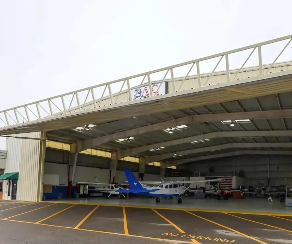 Inside an aircraft hangar with several small airplanes parked, including a blue and white plane in the center, and American flags and an FAA sign visible.