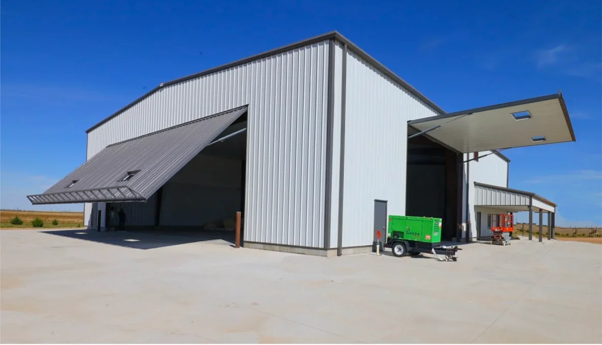 A large metal industrial warehouse building with multiple roll-up doors and extended awnings, situated on a concrete lot under a clear blue sky.
