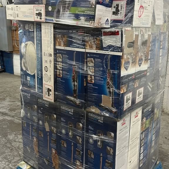 Stack of boxed Bissell vacuum cleaners and other home appliances wrapped in plastic on a wooden cart in a store aisle.