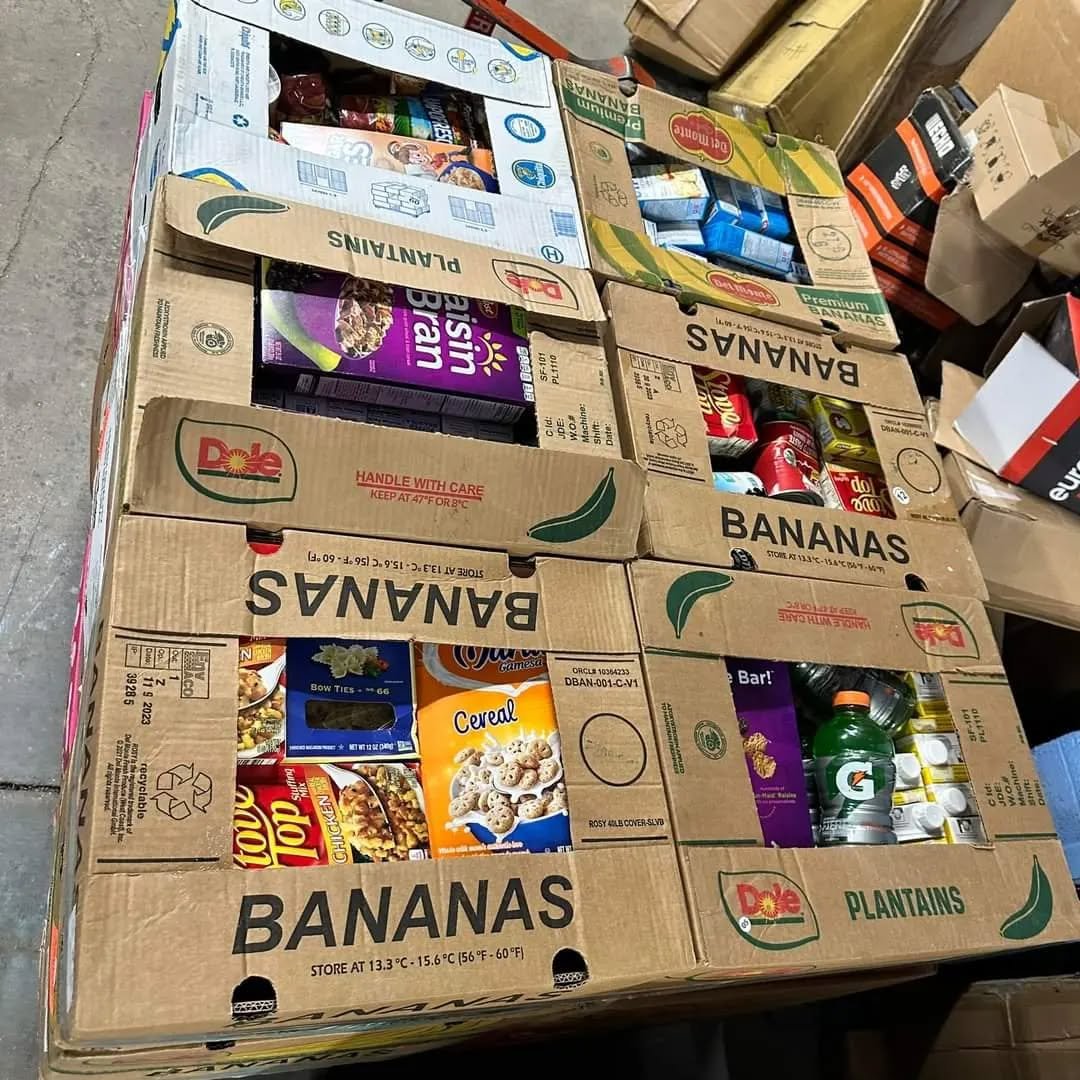 Stacked boxes of groceries including bananas, cereal, chips, snacks, and canned drinks in a warehouse or store.