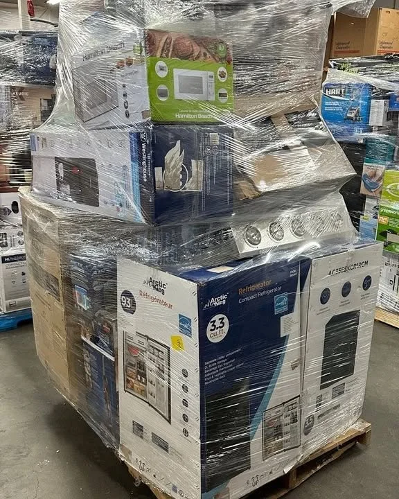 Pallet wrapped in plastic containing various appliances, including a refrigerator, microwave, and other boxed items, in a warehouse setting.