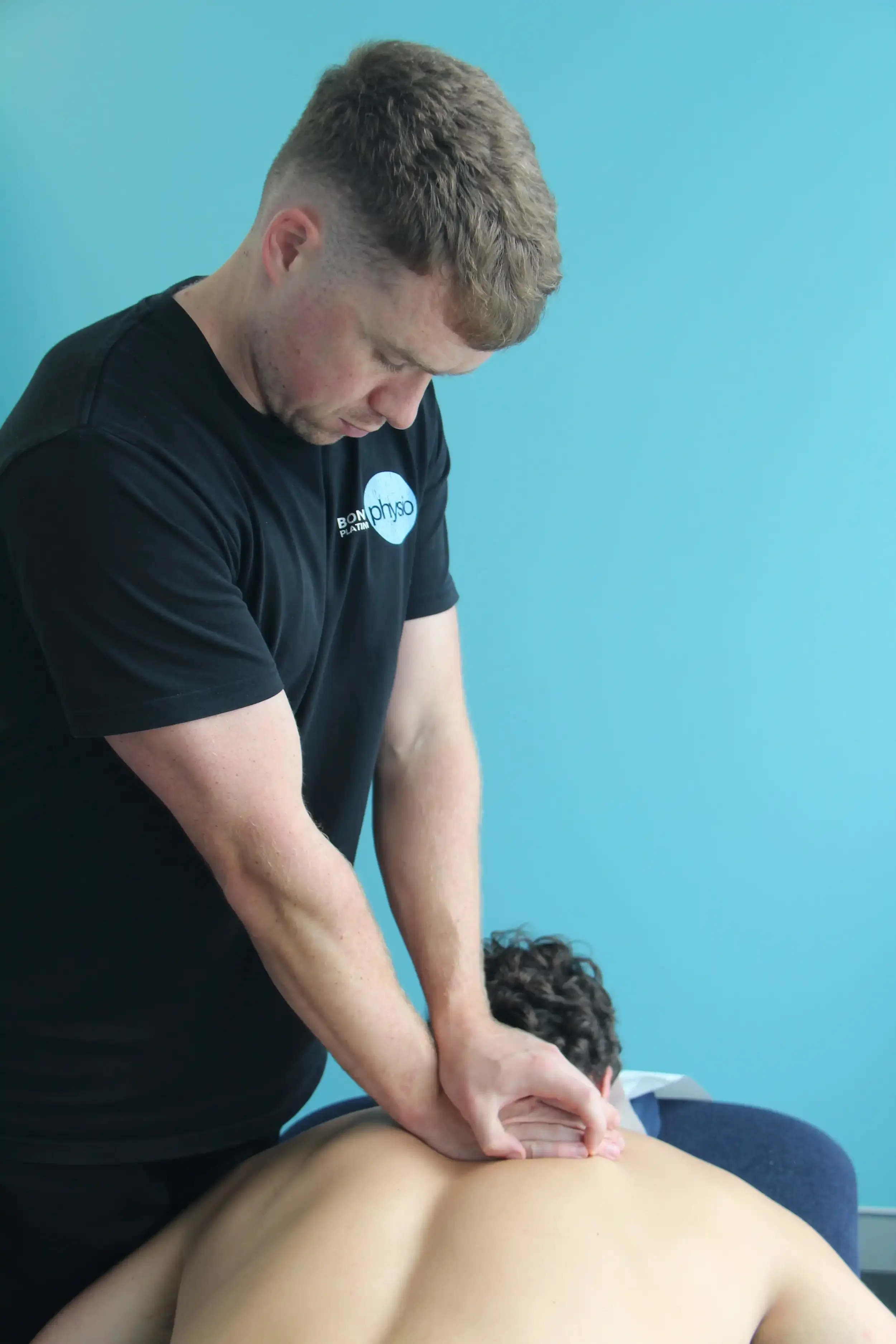 A physiotherapist giving a massage or therapy to a person lying face down, with a blue wall in the background.