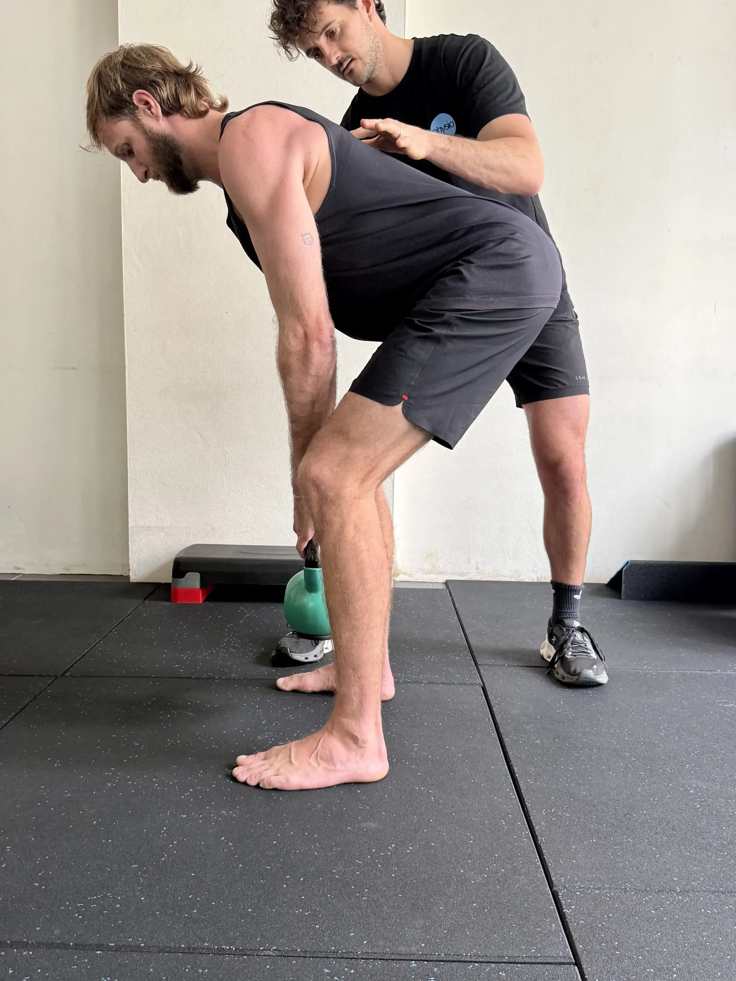 A man is performing a deadlift exercise with a kettlebell while a trainer supervises him. The man is bent over with his hands on his shins, and the trainer is standing behind him, observing and providing guidance. They are in a gym with black rubber flooring.