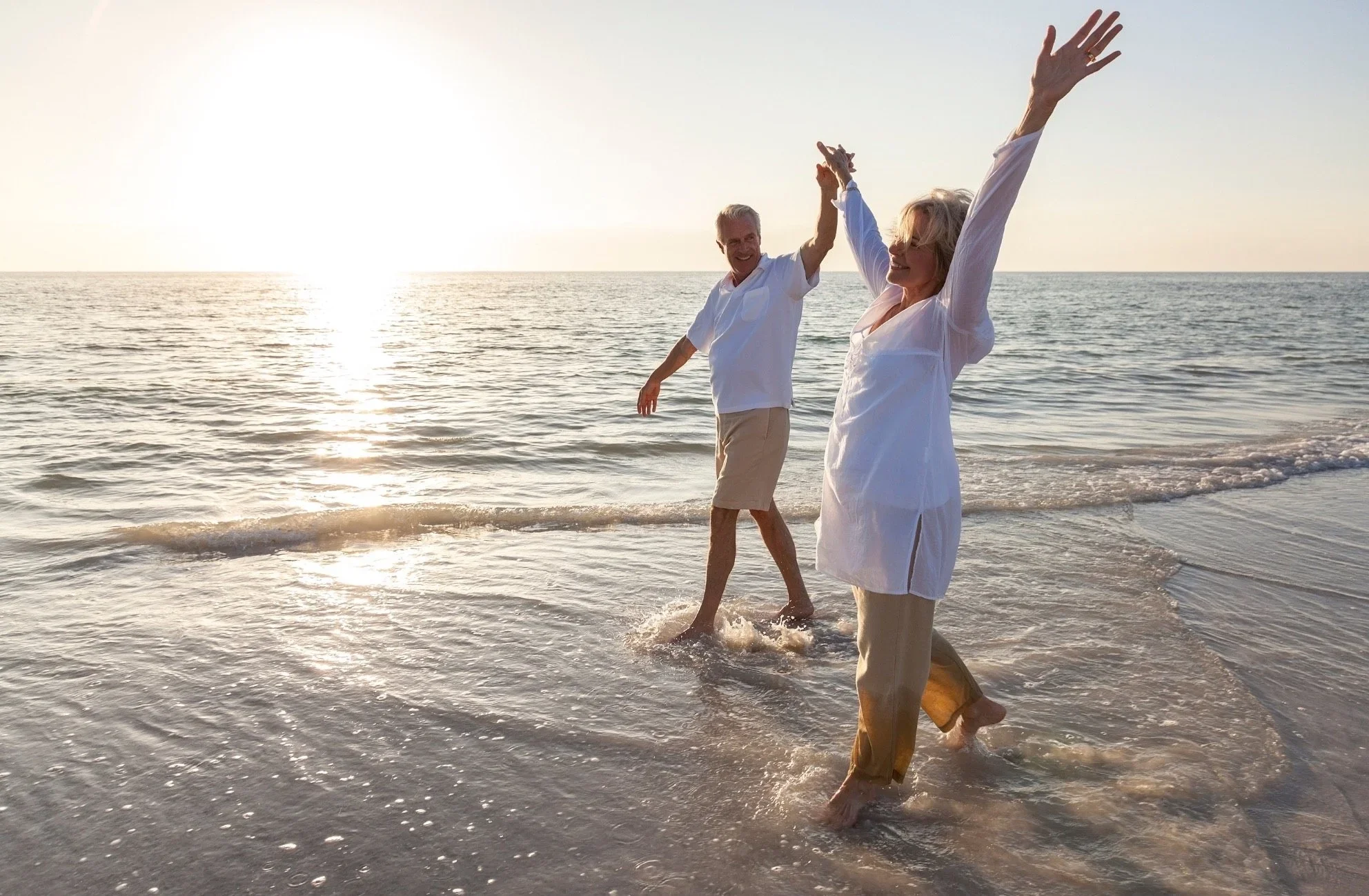 Two elderly people, a man and a woman, joyfully walking on the beach at sunset, holding hands and smiling.