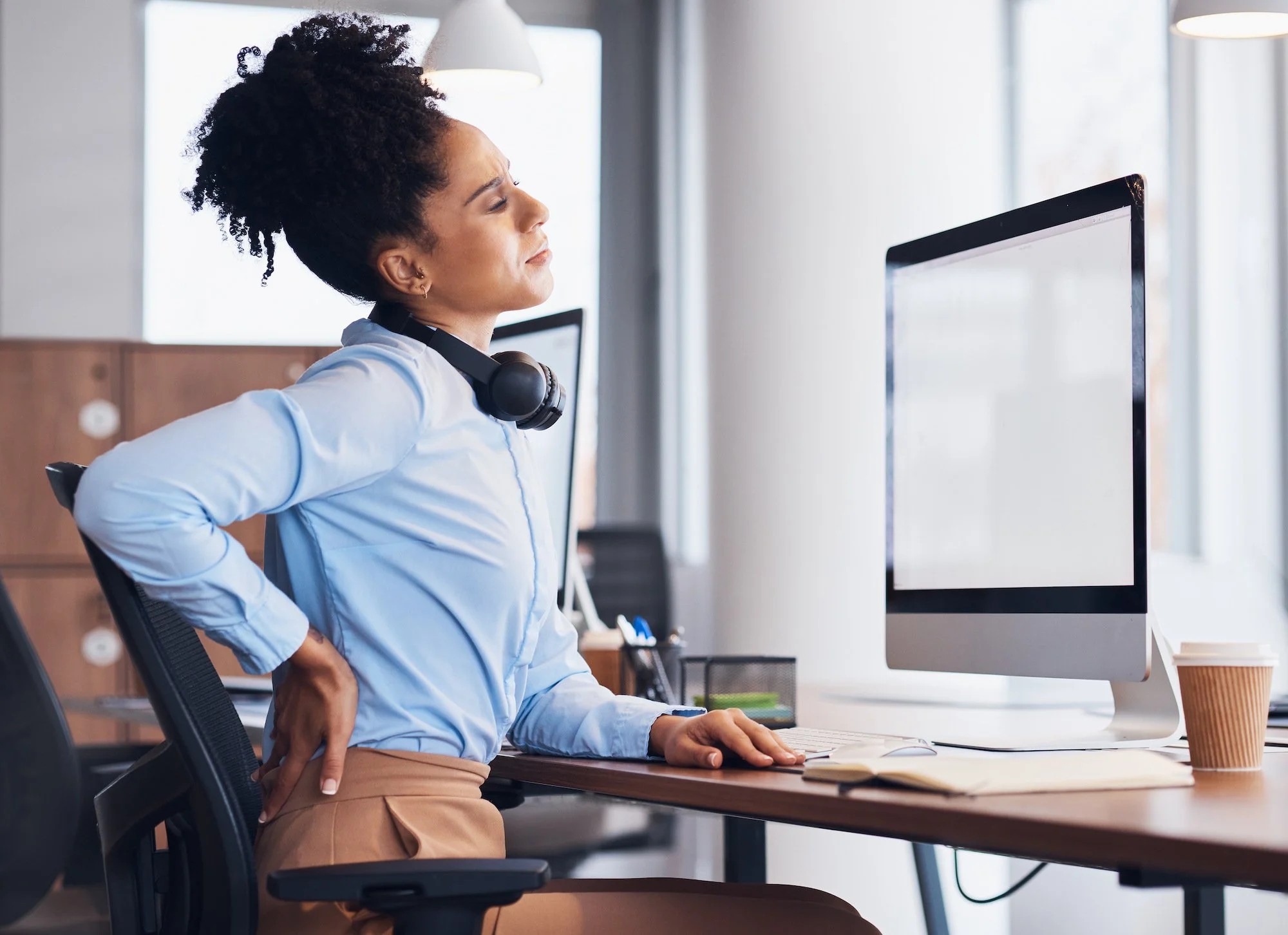 A woman with curly hair sitting at a desk in front of a computer monitor, wearing a blue shirt and beige pants, in a modern office setting.