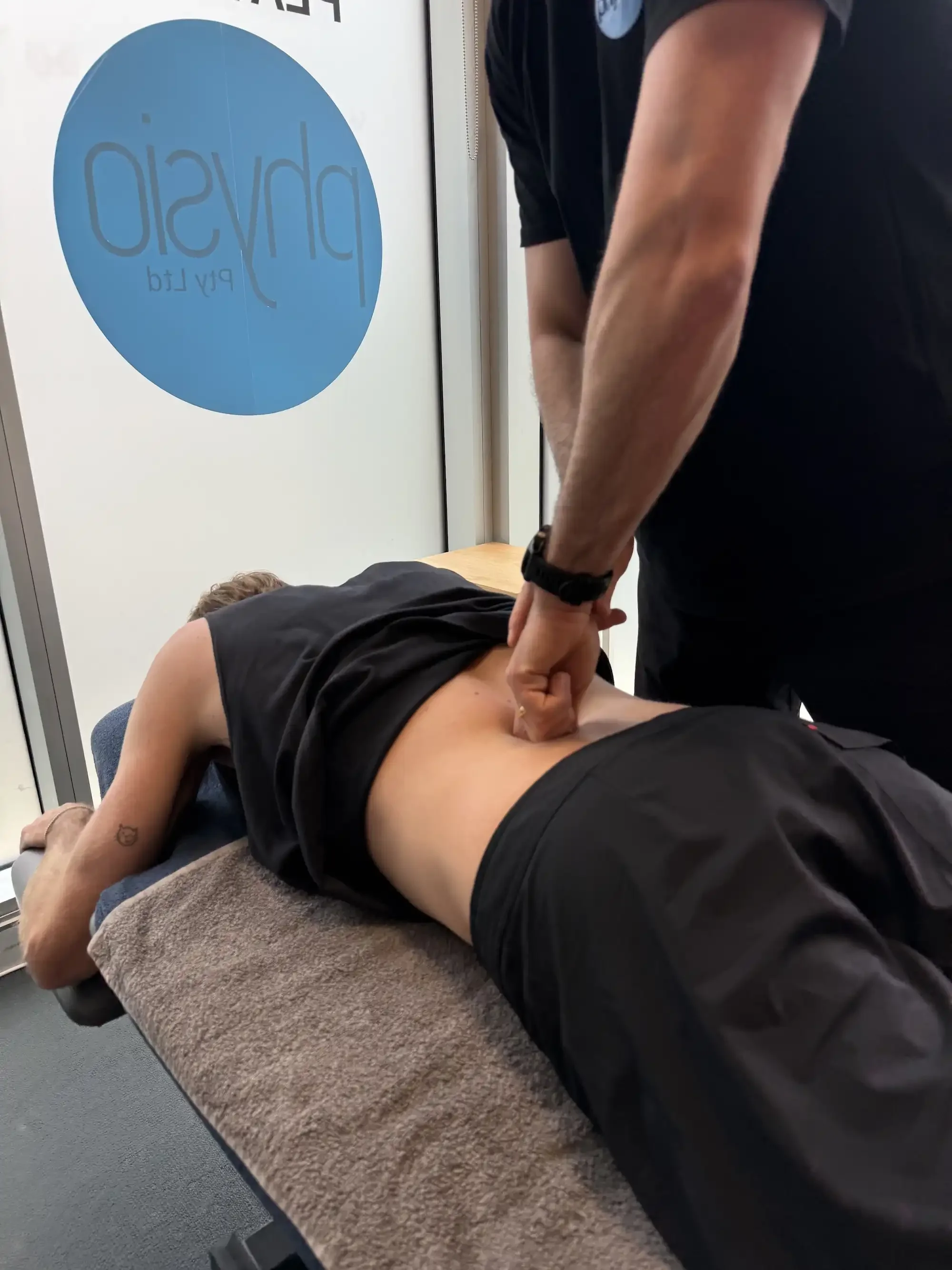 A person lying face down on a padded table or bed while another person performs a procedure or massage on their back. The setting appears to be a clinic or massage therapy room.