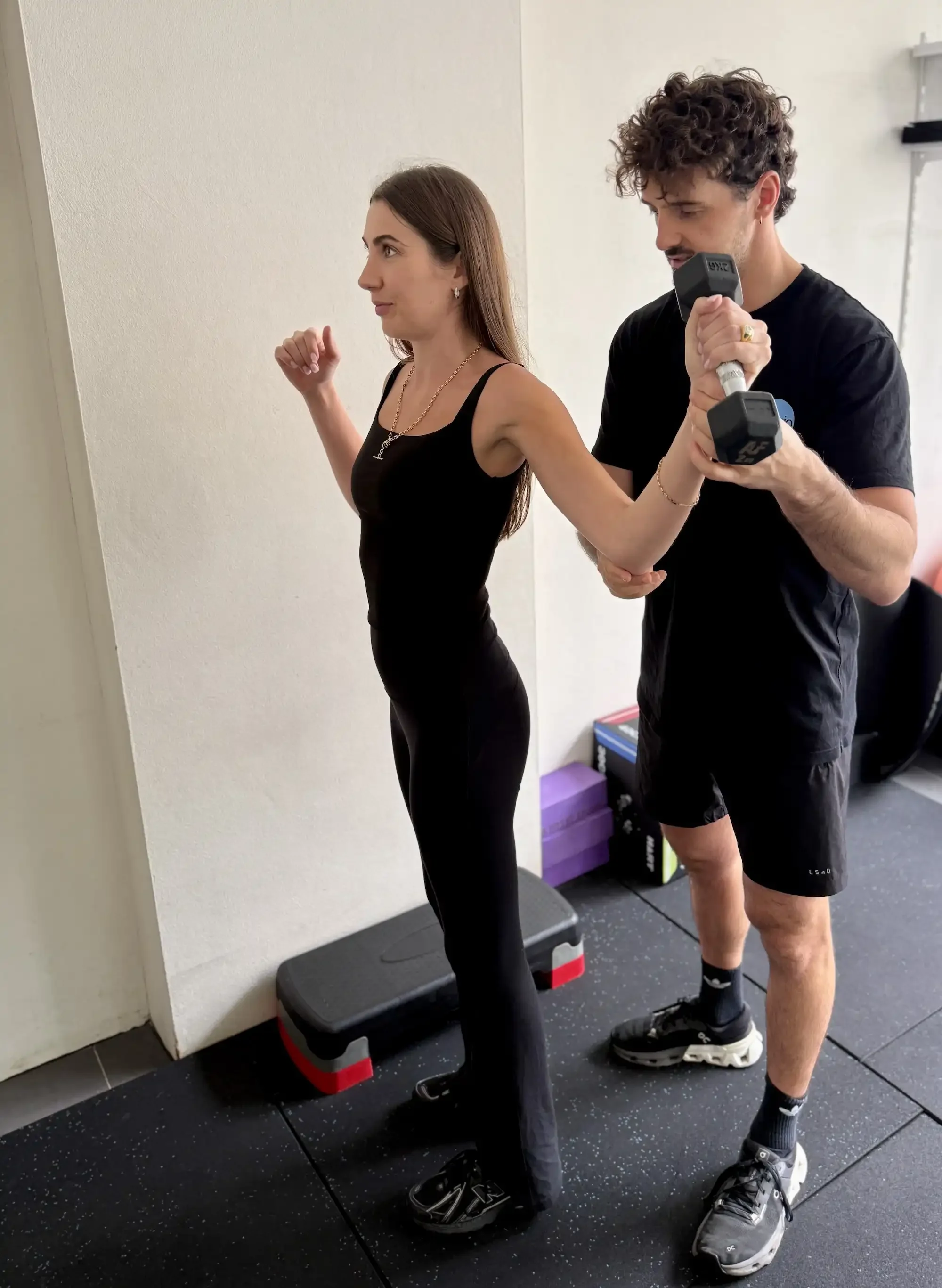 A woman in black workout clothes doing a step exercise in a gym while being guided by a man holding dumbbells.