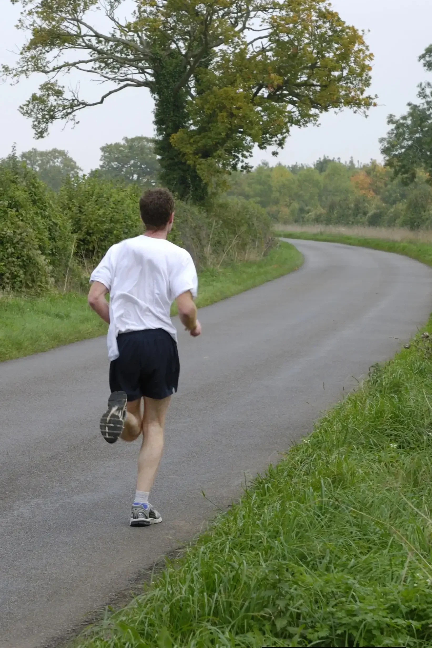 A man running on a paved country road surrounded by green grass and trees on a cloudy day.