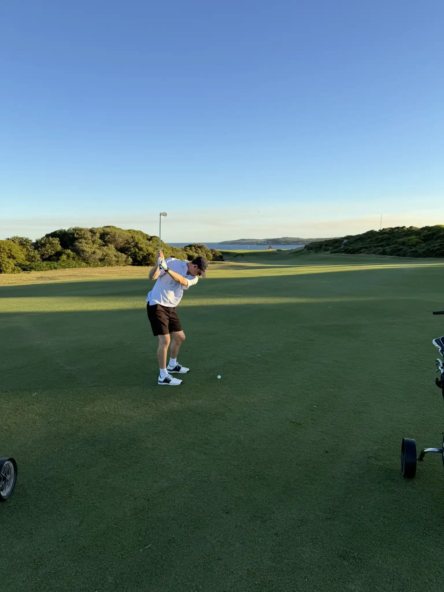 A man in a white shirt and black shorts prepares to hit a golf ball on a green golf course during late afternoon or early evening.