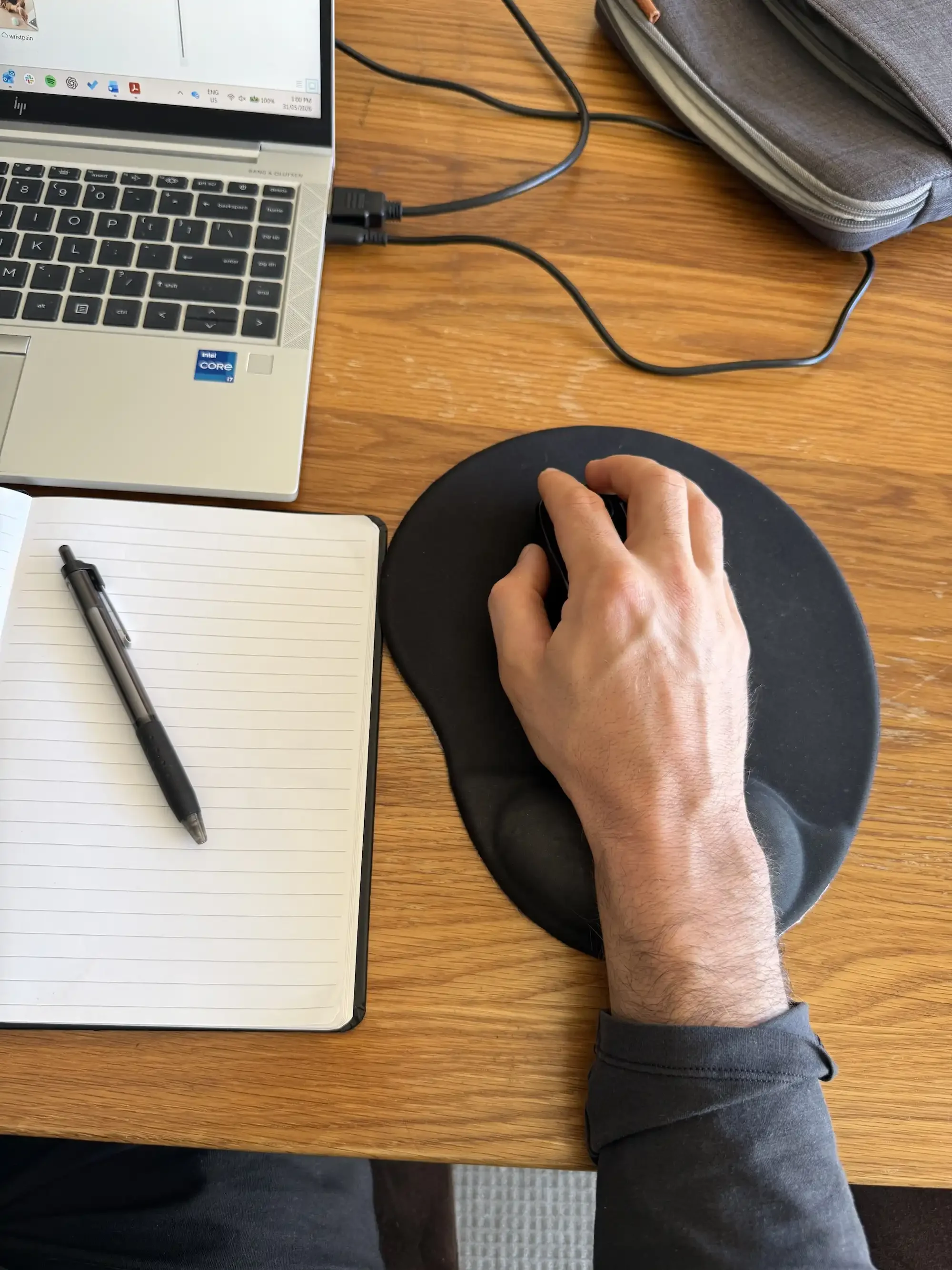 A hand resting on a computer mouse on a black mouse pad, on a wooden desk with a laptop, an open notebook with a black pen, and a gray backpack nearby.