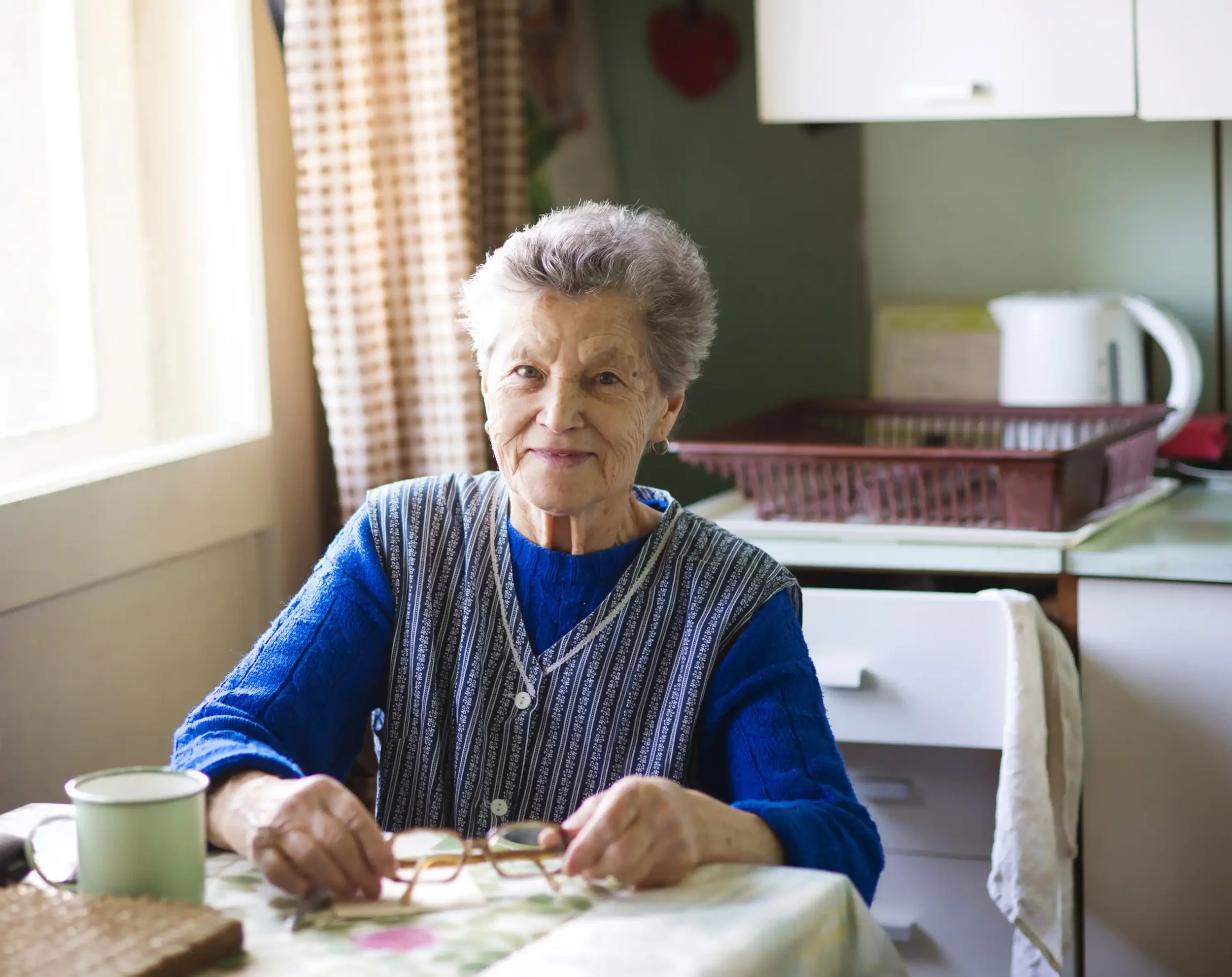 An elderly woman with white hair and a gentle smile sitting at a table in a cozy room, wearing a blue sweater and a patterned apron, with two mugs and a pair of glasses in front of her.