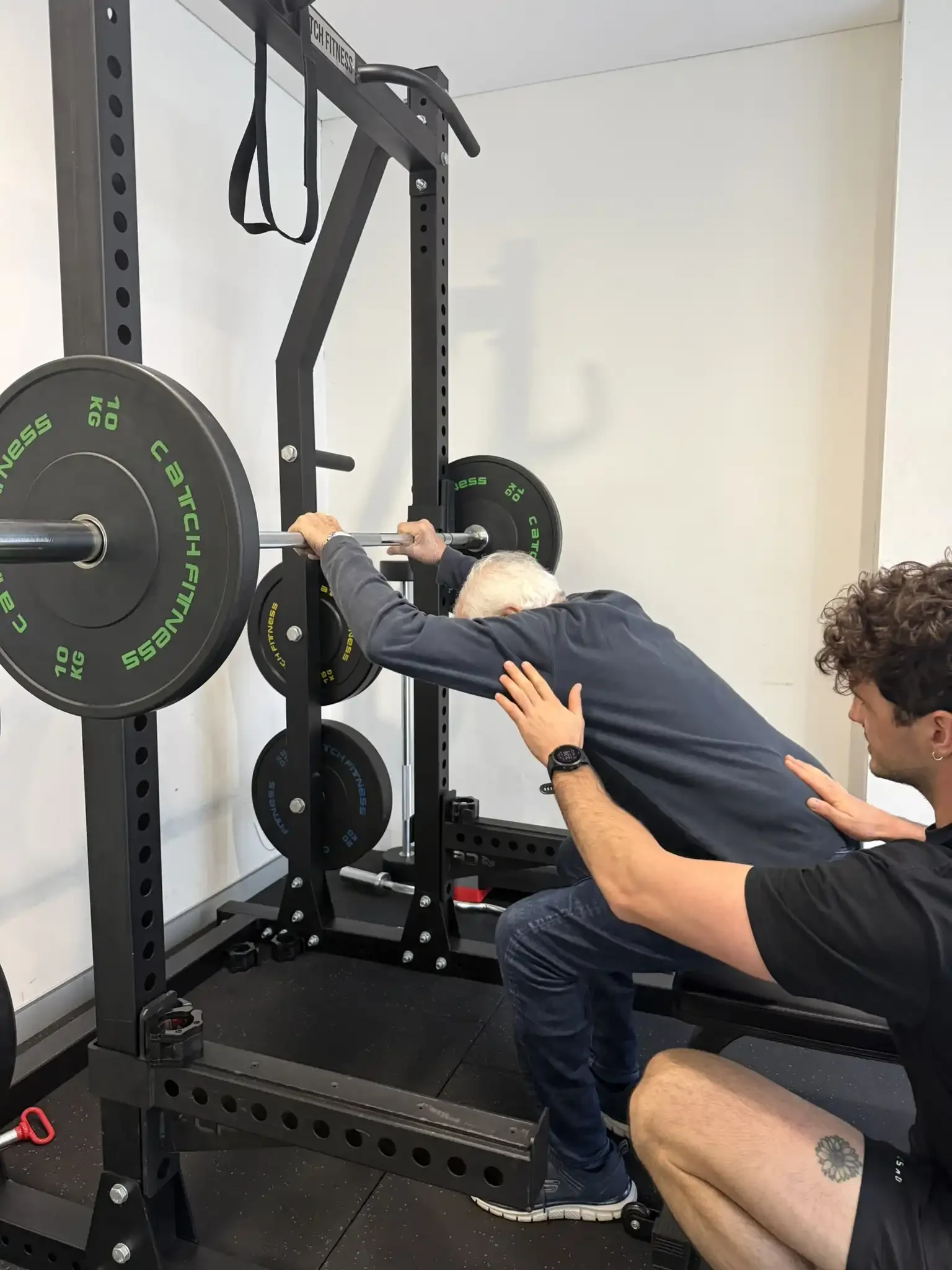 A person is performing a squat exercise with a barbell on their shoulders in a gym. The barbell has weight plates labeled 10 kg, and there are other weights on the rack nearby. Another person is spotting or coaching, with a hand on the lifter's back for support.