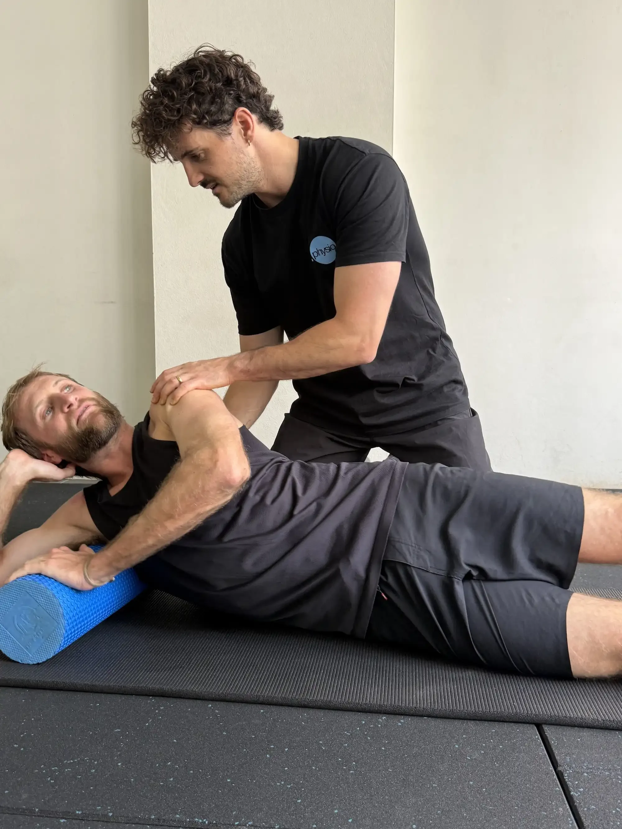 A fitness instructor assisting a man with a leg exercise on a yoga mat. The man is lying on his side with his head supported by his hand, and the instructor is holding his leg and providing guidance.