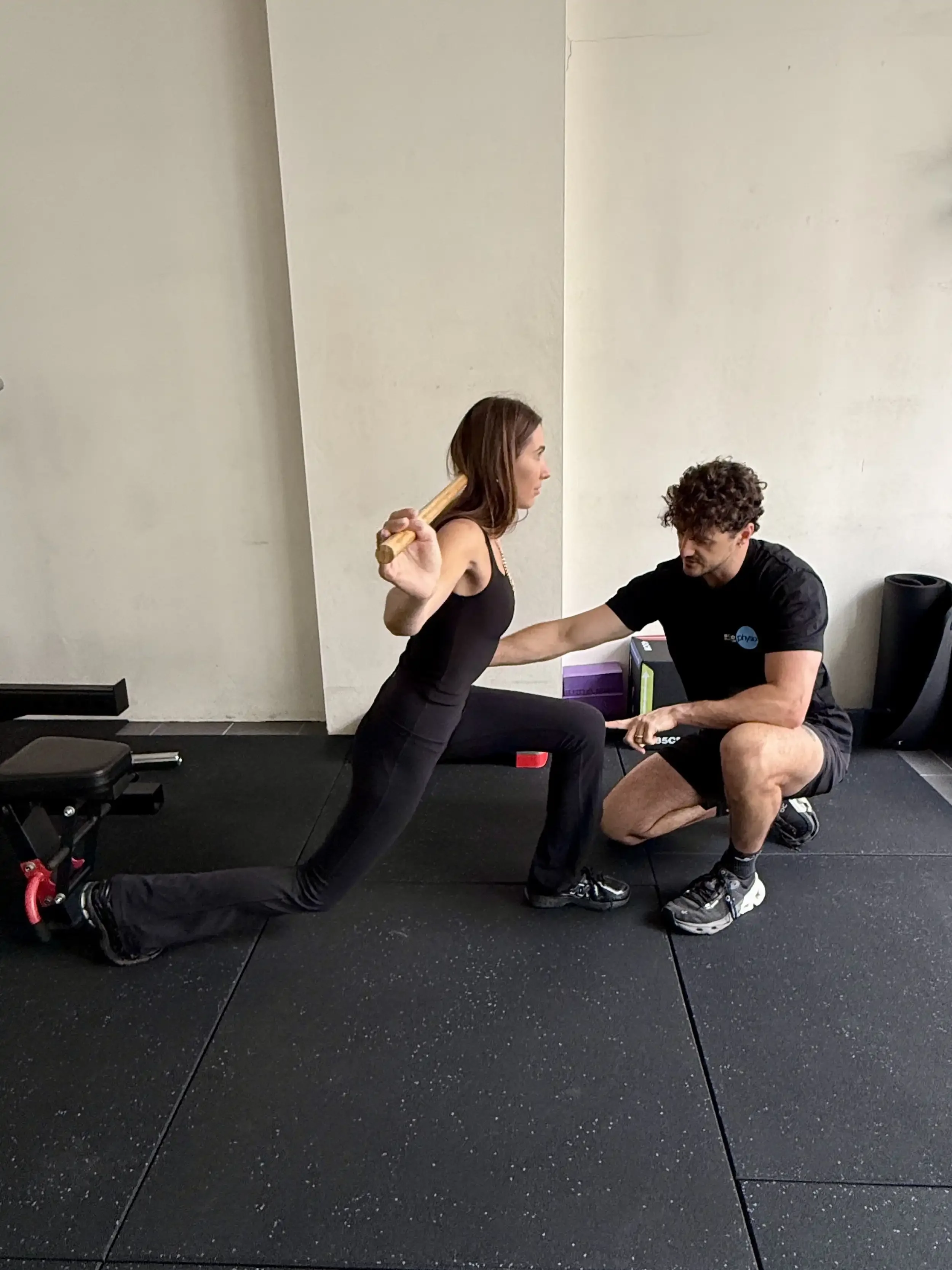 A woman performing a lunge exercise with a wooden stick supported by a trainer in a gym. The trainer is crouching and holding the woman’s hand for assistance.
