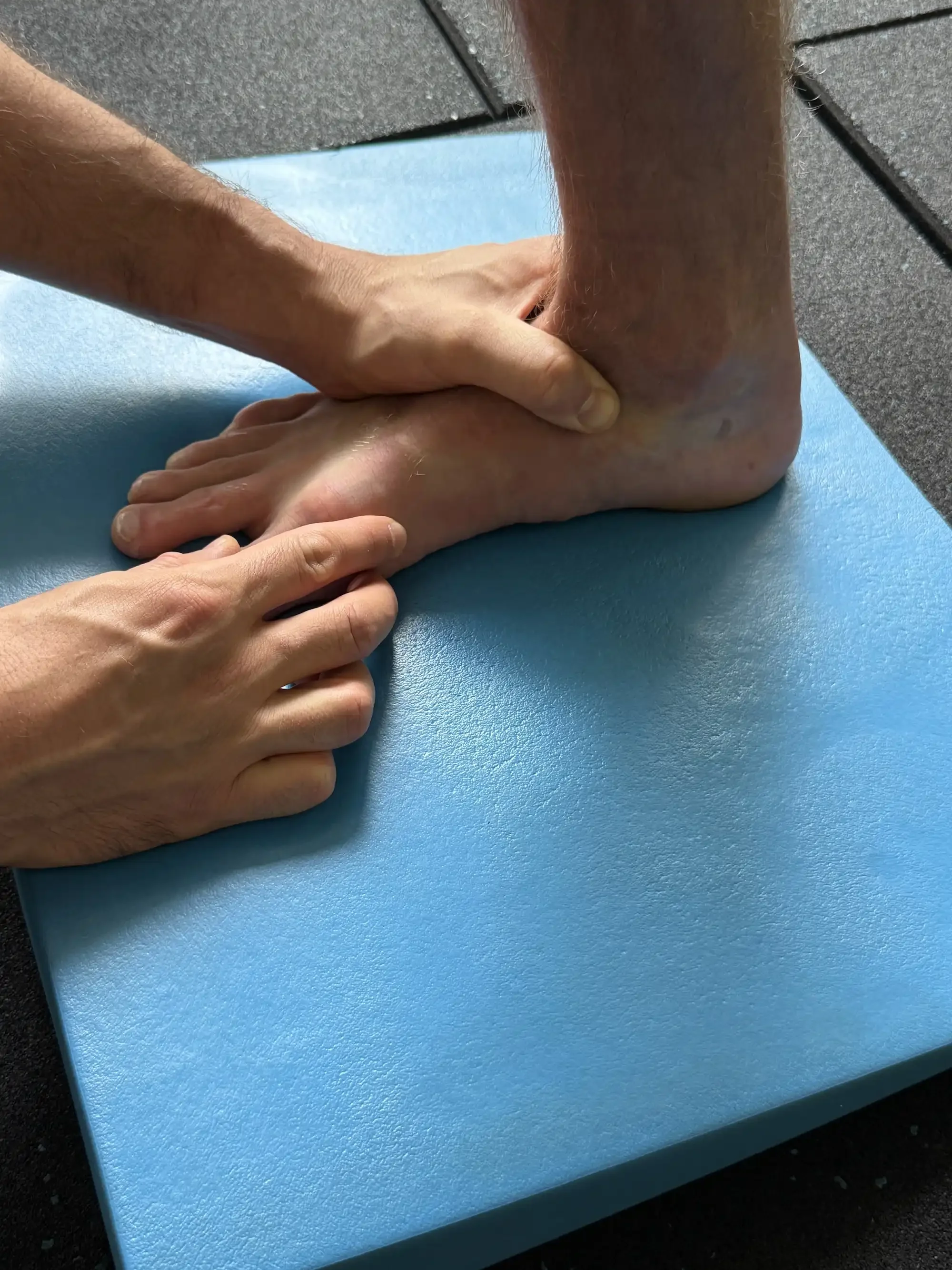 A person performing a physical therapy or stretching exercise by holding their ankle with one hand and supporting their foot with the other on a blue mat.