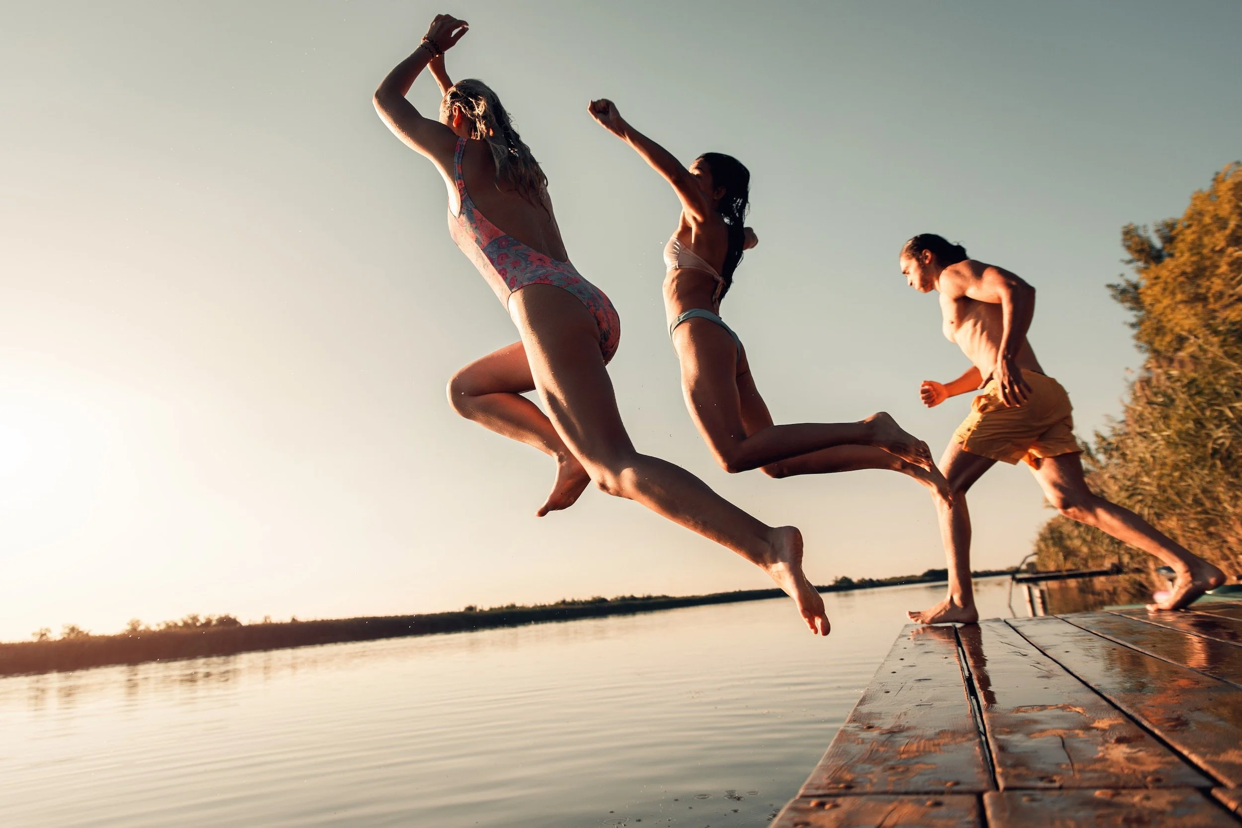 Three people jumping off a dock into the water at sunset or sunrise, with trees in the background.