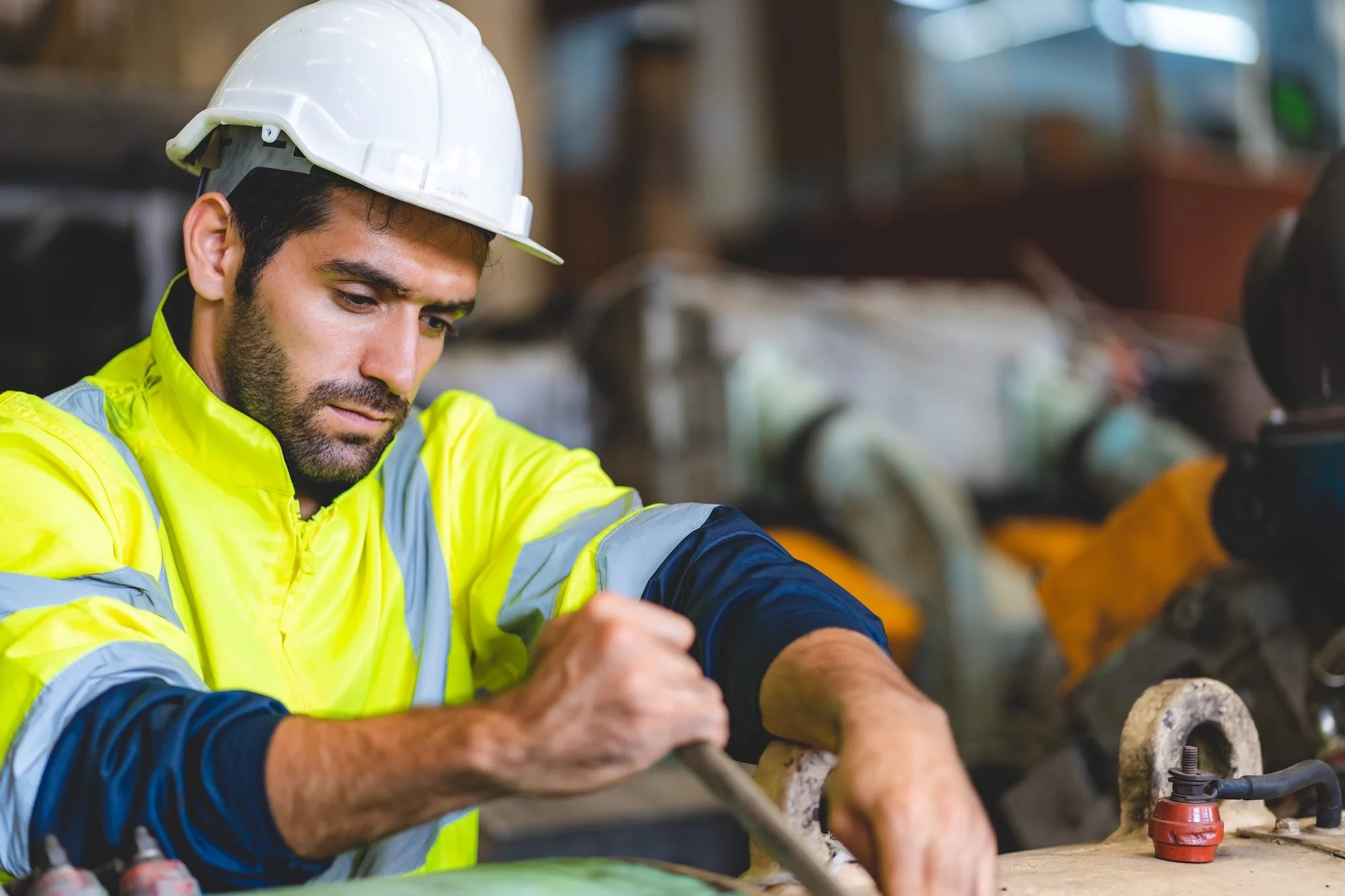 A man wearing a white hard hat and a bright yellow safety jacket is working on a piece of equipment in an industrial or workshop setting.