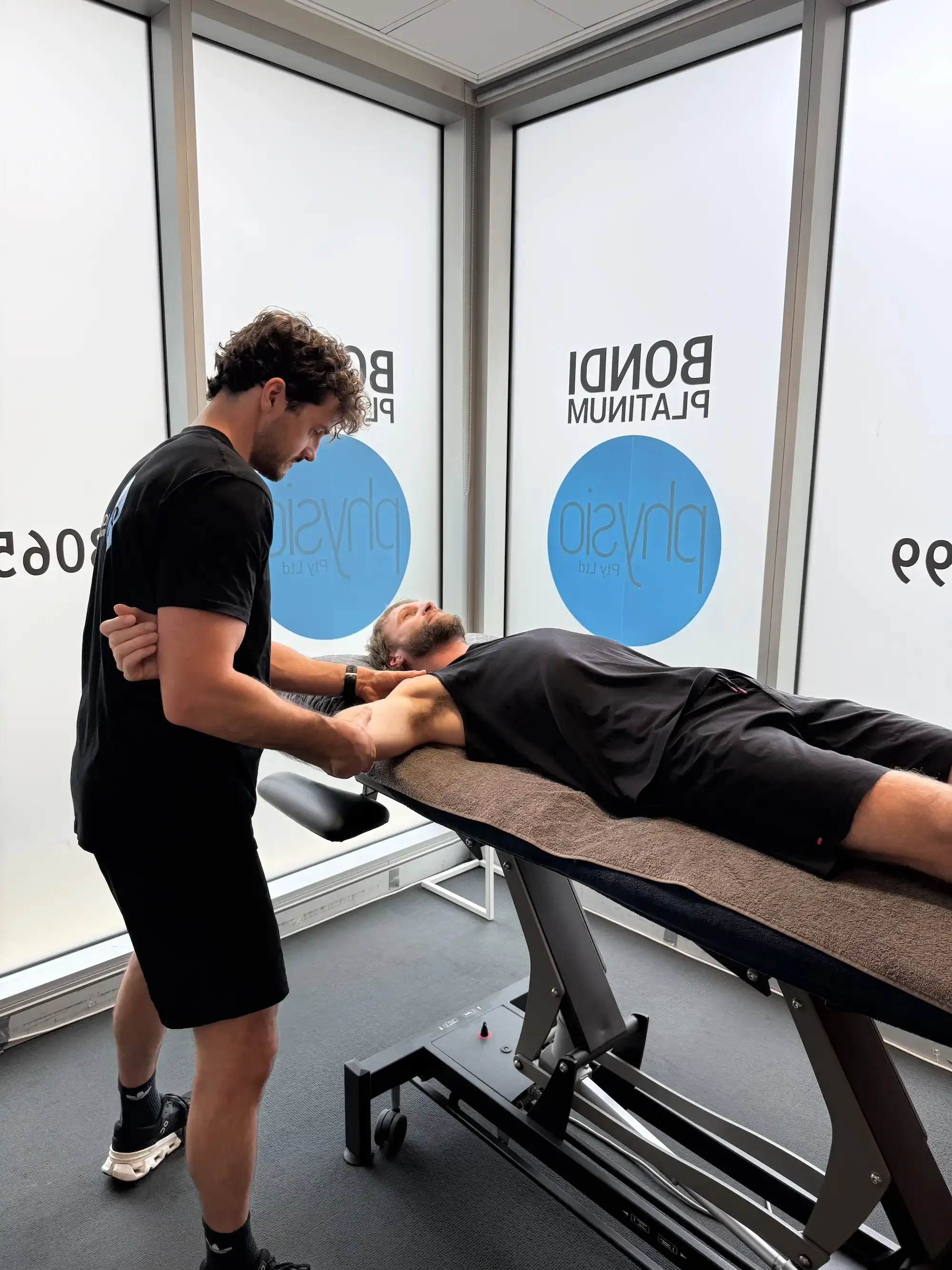 Man lying on a therapy table receiving treatment from another man standing beside him, inside a room with large windows and signage reading 'BOND PLATINUM' in the background.