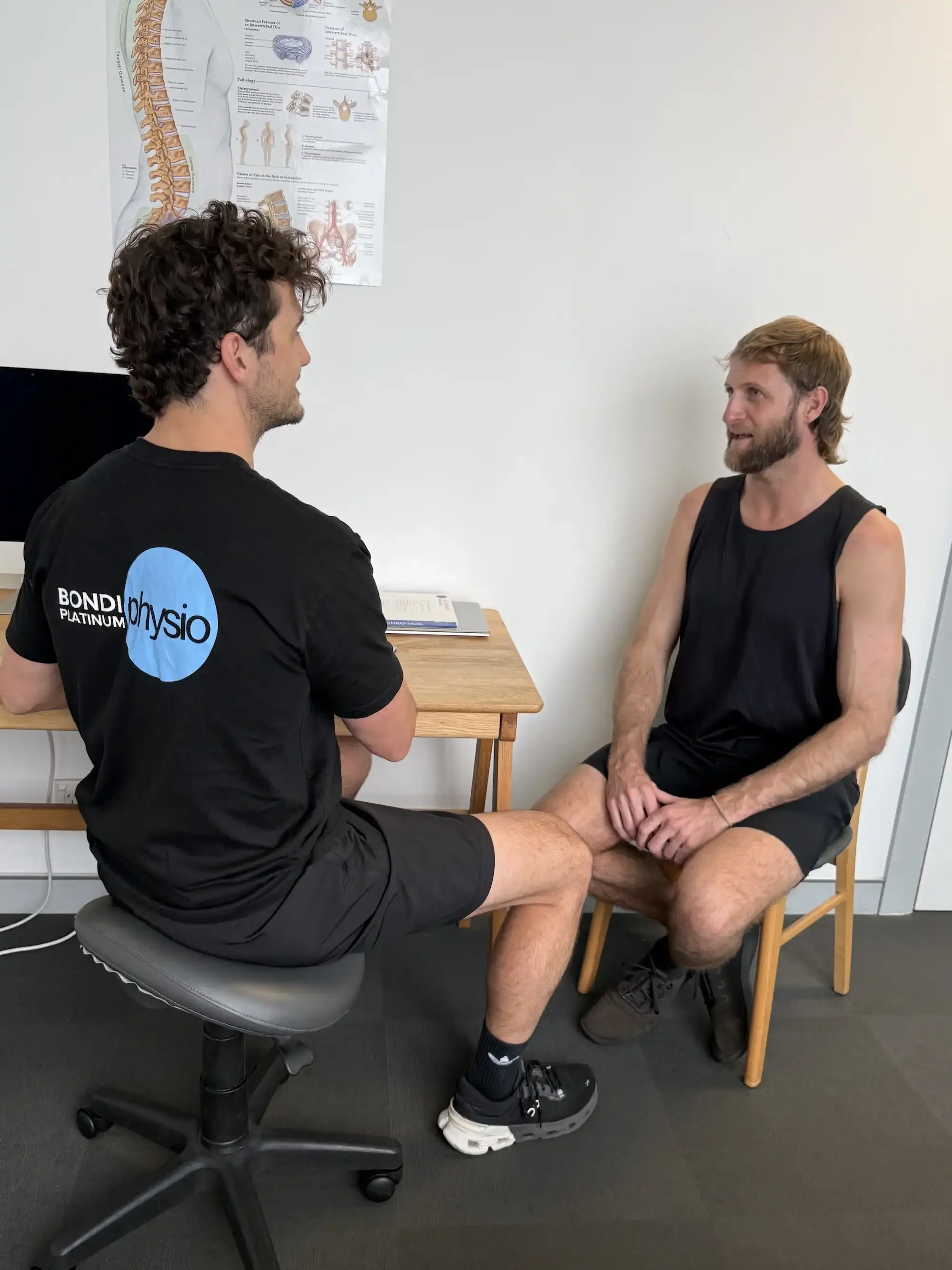 A physical therapist consulting with a male patient during a physical examination in a clinical setting. The patient is sitting on a chair, wearing a black tank top and shorts. The therapist is sitting on a stool, wearing a black shirt with a blue logo and black shorts, holding the patient's leg in a clinical assessment. There is a medical poster about the spine on the wall behind them.