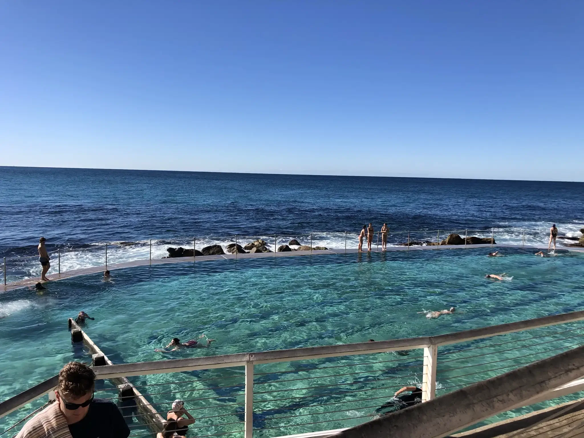 A scene at a beach with a swimming area separated by a barrier from the open ocean. Several people are swimming and relaxing, the ocean and a clear blue sky are visible in the background.