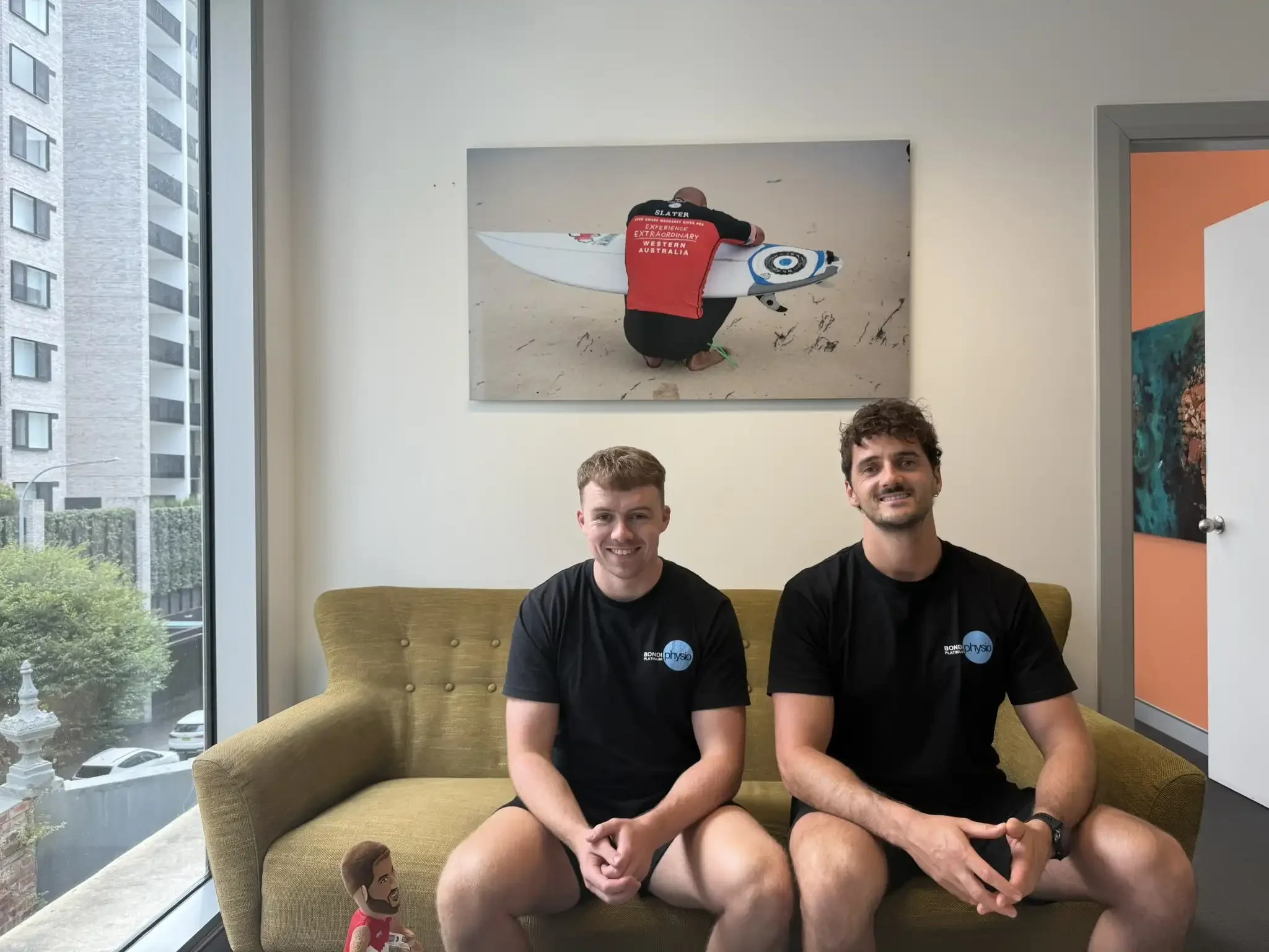 Two men sitting on a yellow sofa in a bright room with large windows, smiling at the camera. One man has curly dark hair, and the other has short brown hair. Both are wearing black t-shirts with a logo that says 'physio'.