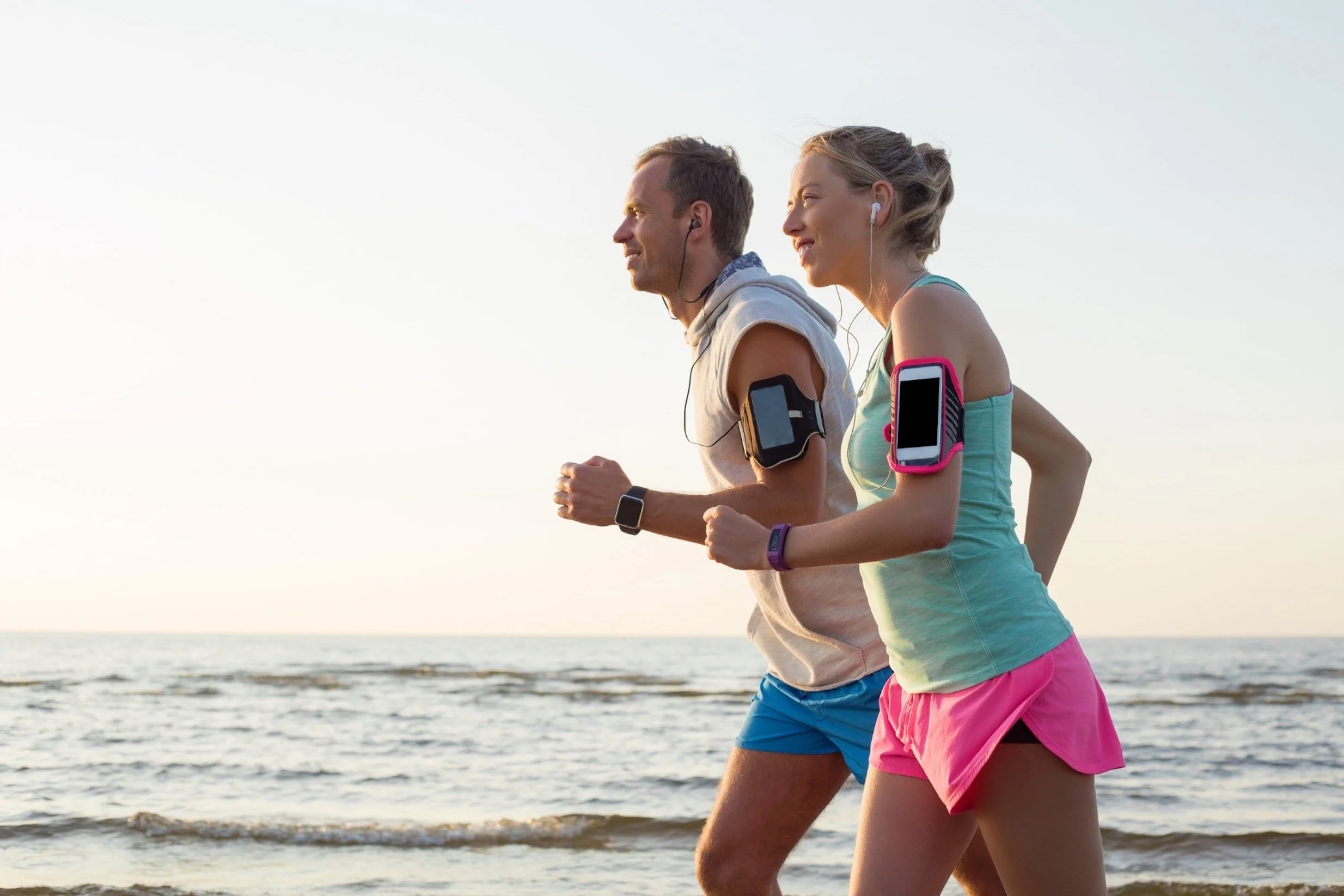 Two people jogging along the beach during sunset, wearing athletic clothing and earbuds, with smartphones in armbands.