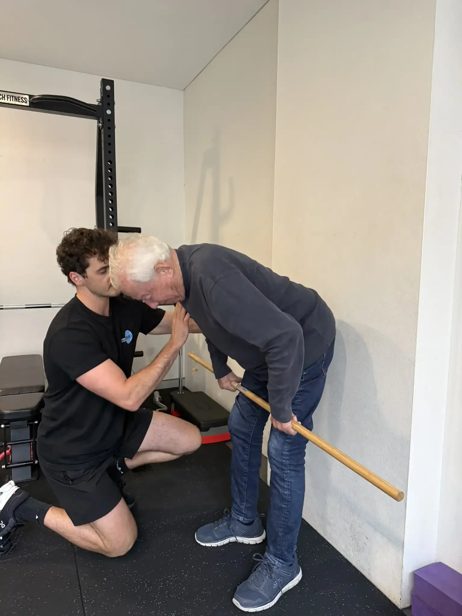 A young man kneeling and lifting a leg while an older man with white hair aids him, holding a wooden staff. They are in a gym. The older man appears to be guiding or assisting the exercise.