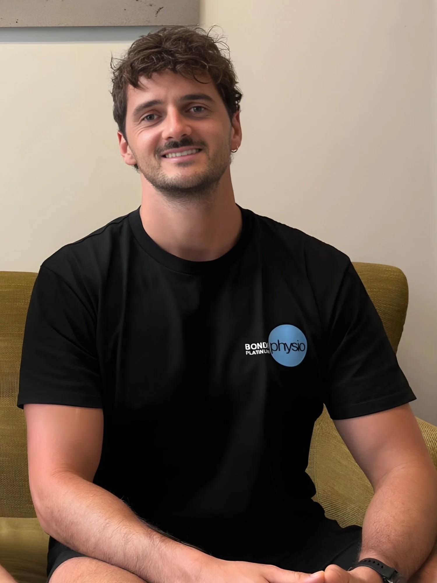 A smiling young man with dark hair wearing a black t-shirt with a logo that says "physio" is sitting on a yellow chair in a room with beige walls.