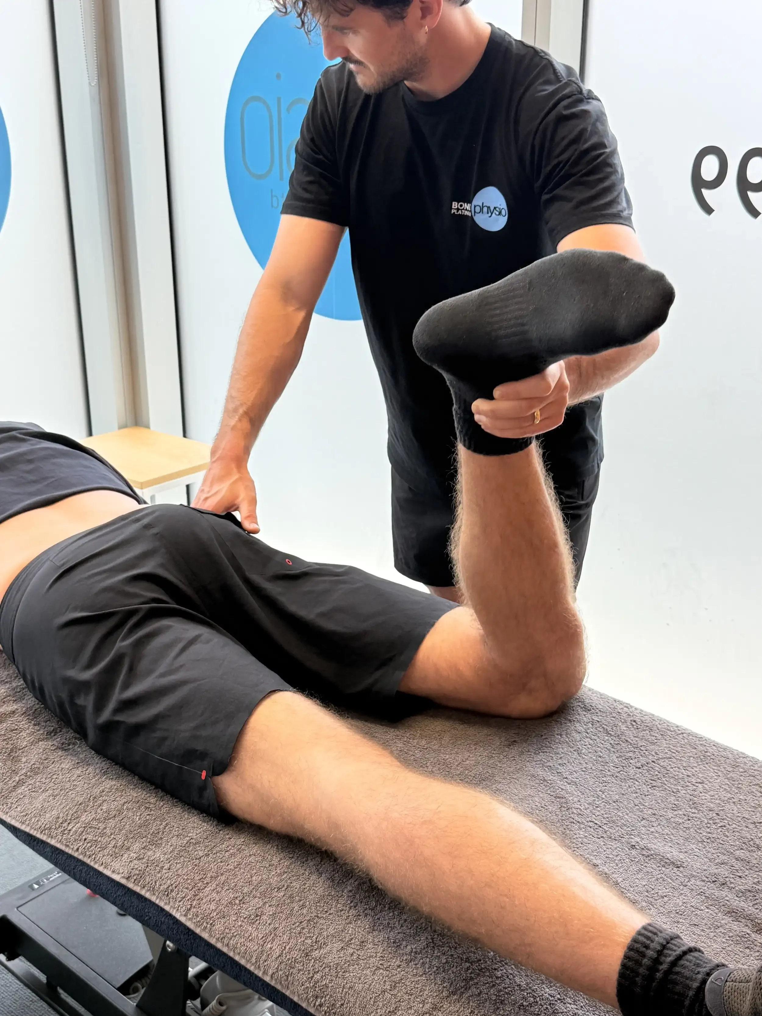 A man lying face down on a massage table with a towel covering his lower body, receiving a physical therapy or chiropractic treatment from a therapist. The therapist is standing next to the table, holding the man's foot and wearing a black T-shirt with a logo.