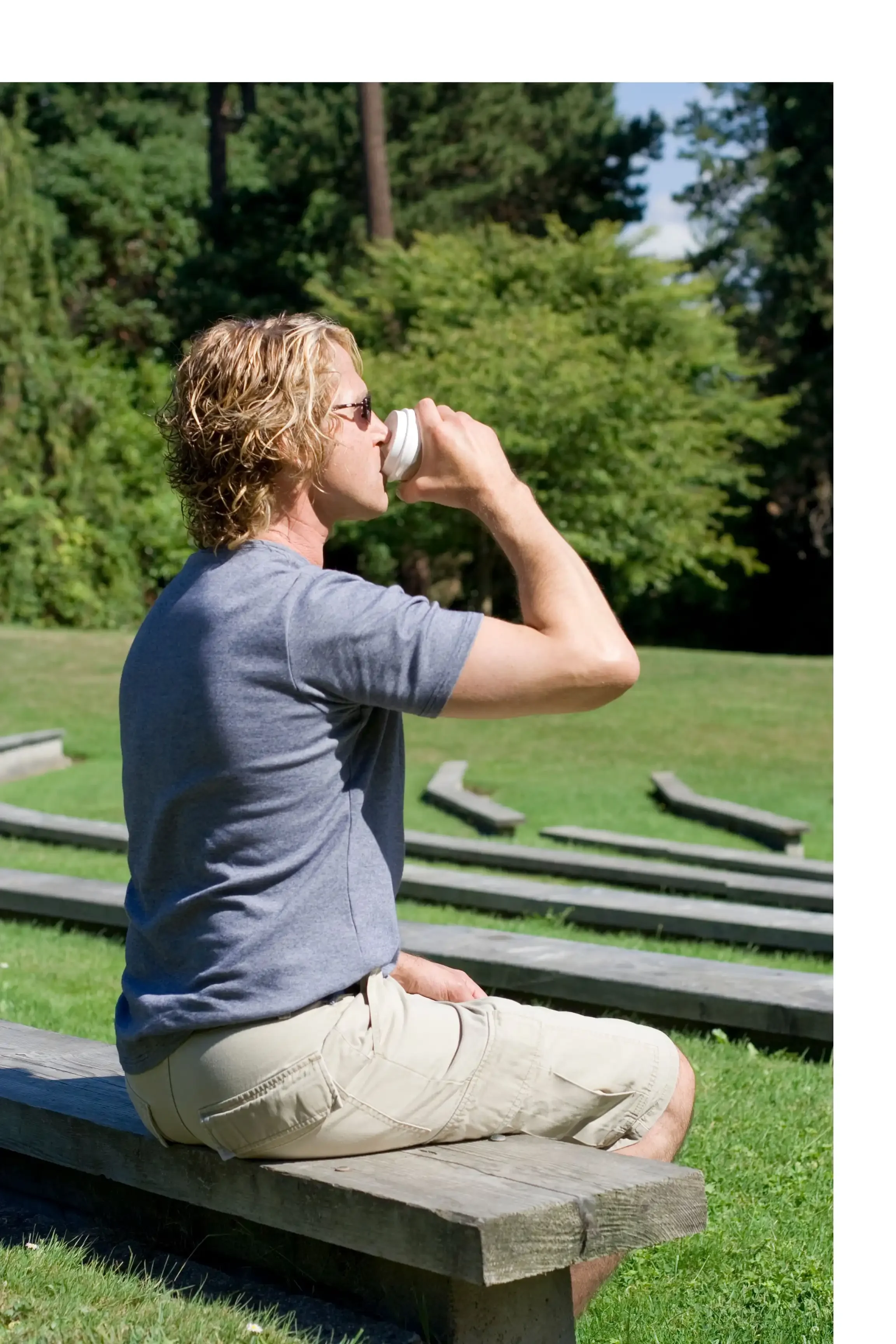 A man with curly blonde hair, wearing glasses, a gray t-shirt, and khaki shorts, sitting on a wooden bench outdoors, drinking from a disposable cup with a lid, with green trees and a grassy area in the background.