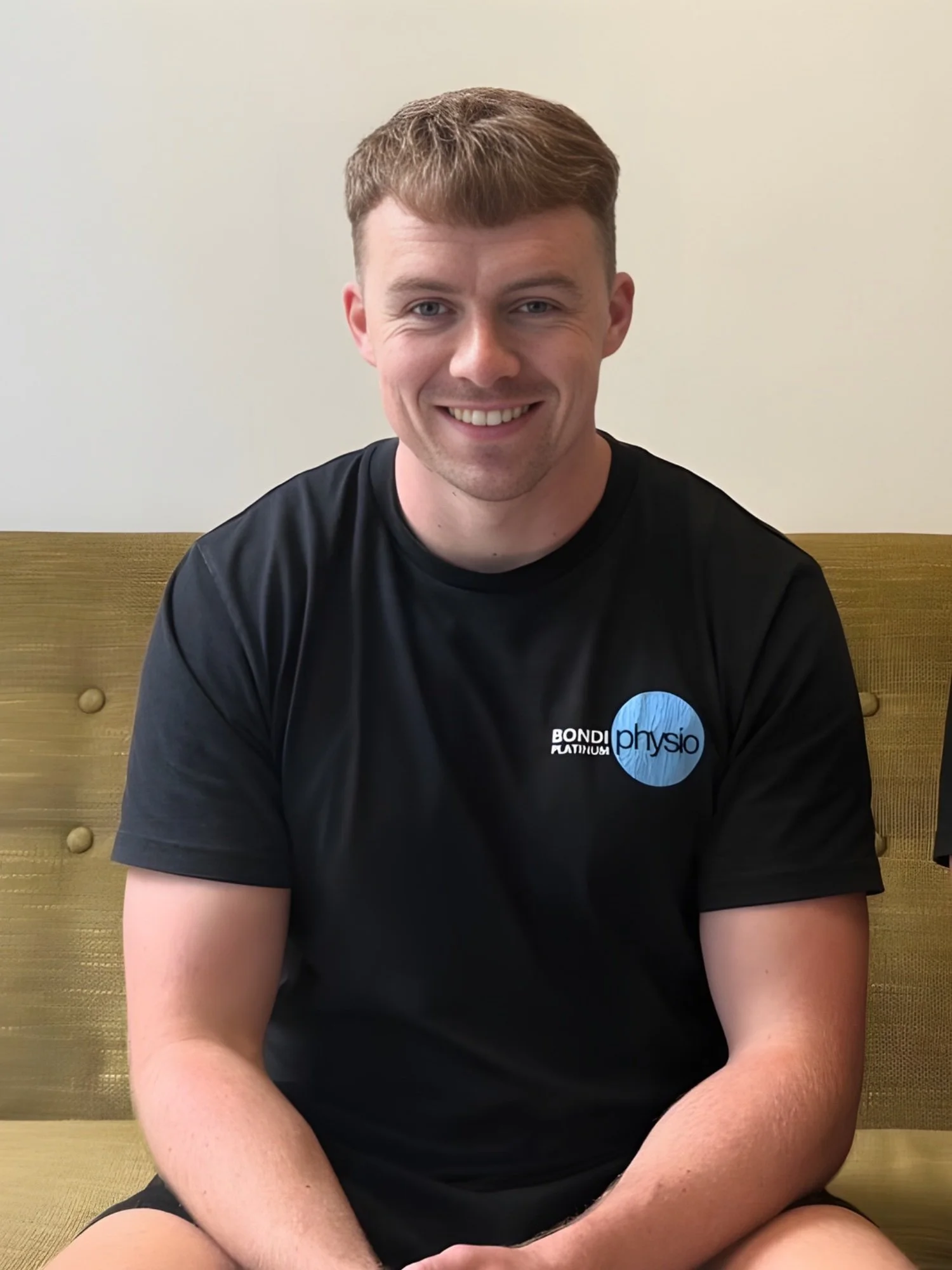 A smiling young man with short brown hair and blue eyes, sitting against a wooden bench with a plain beige wall behind him, wearing a black t-shirt with a blue and white logo that says 'Bondi Physio'.