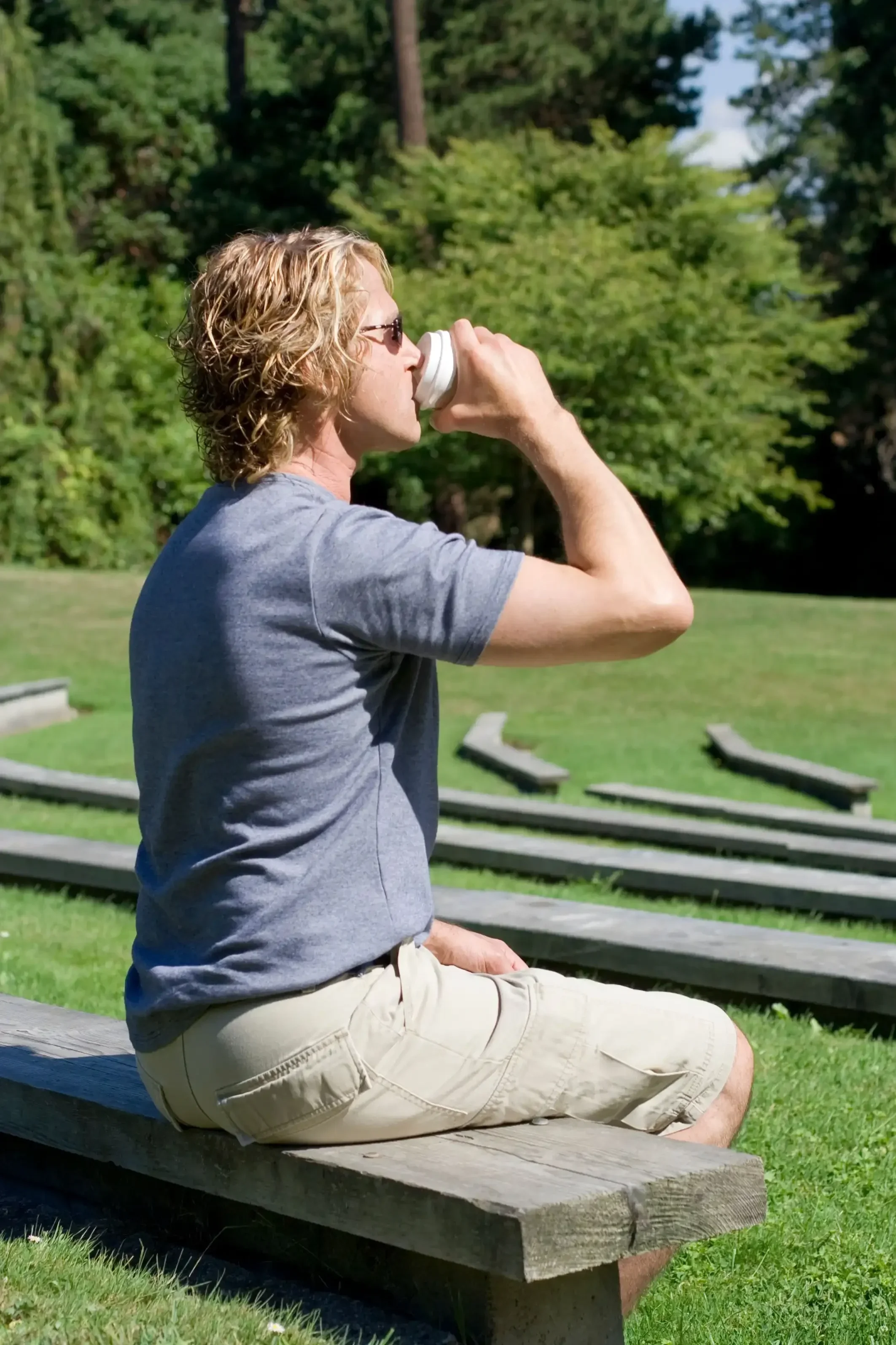A man with wavy blonde hair, wearing sunglasses, a gray t-shirt, and beige shorts, sitting on a wooden bench outdoors, drinking from a white mug, with green trees and a grassy area in the background.