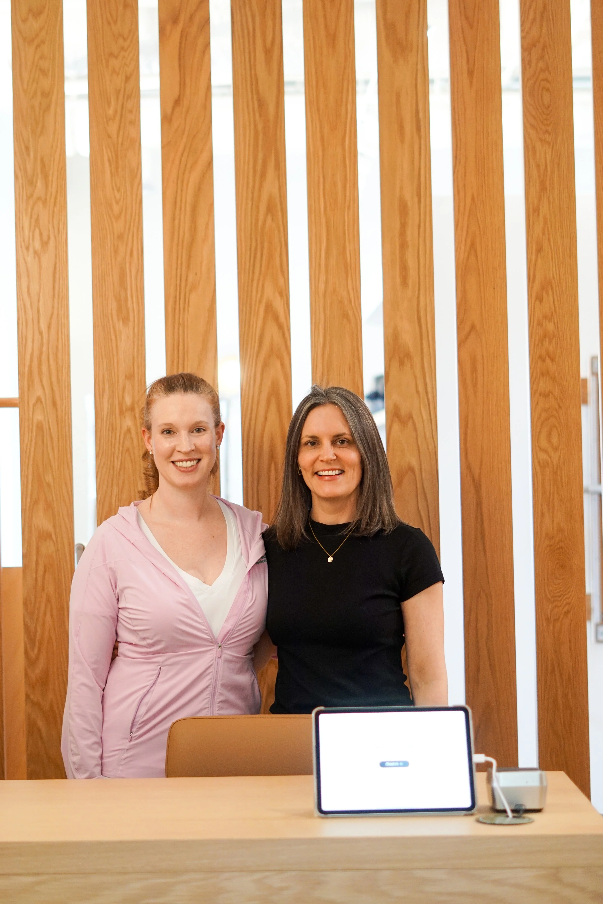 Two women standing behind a reception counter with a tablet and card reader, smiling to welcome someone into their space.
