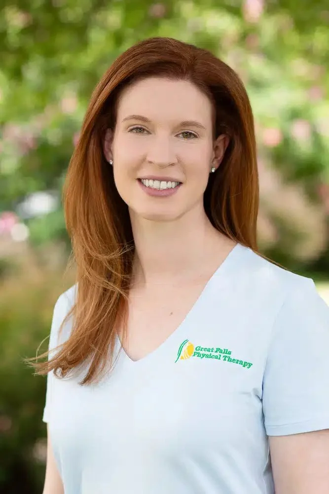 A woman with long red hair smiling outdoors, wearing a white shirt with a logo that says Great Falls Physical Therapy, in front of a green, blurred background.