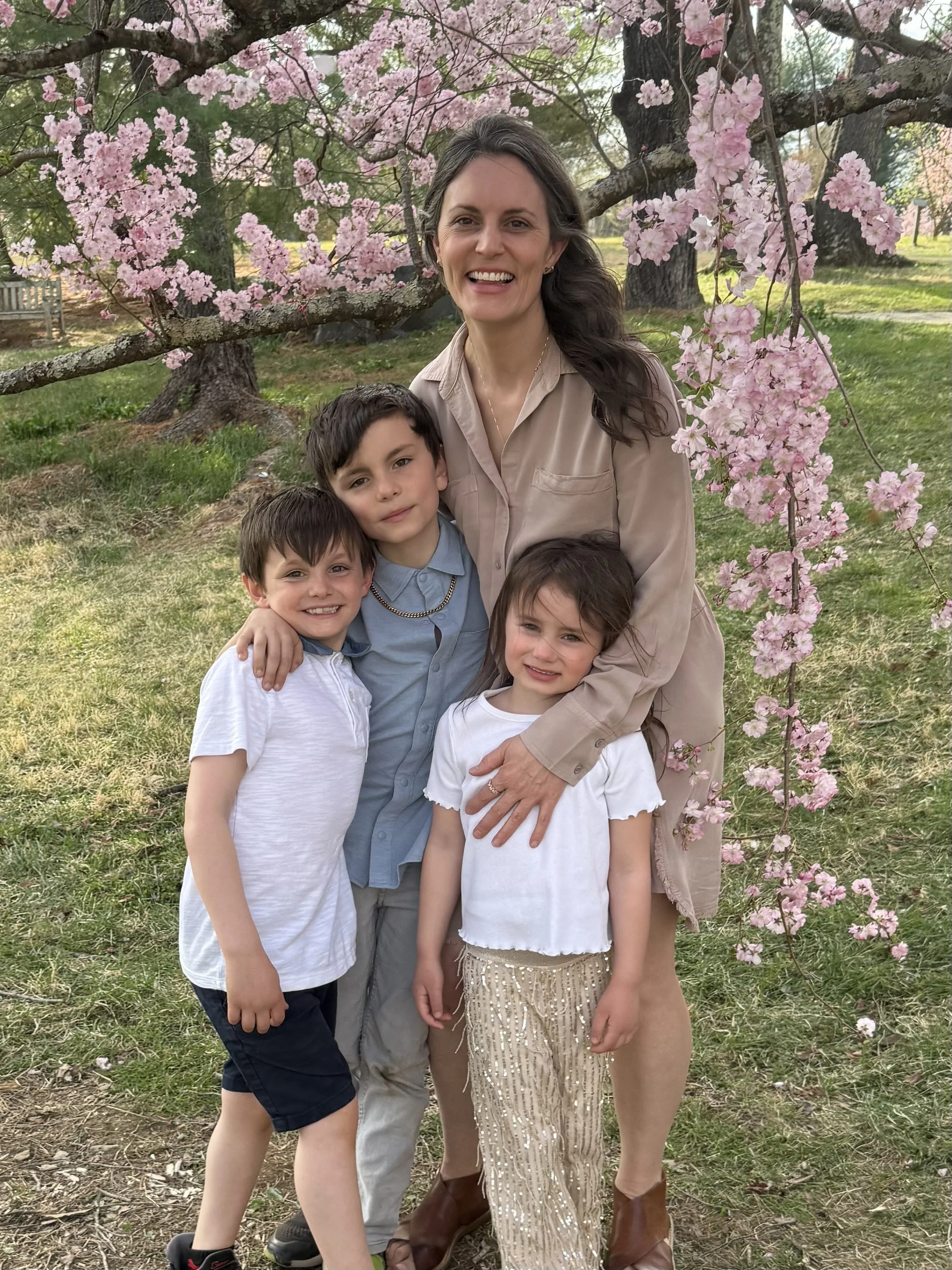 A woman with three children standing outdoors under a blooming pink cherry blossom tree, smiling and hugging them.